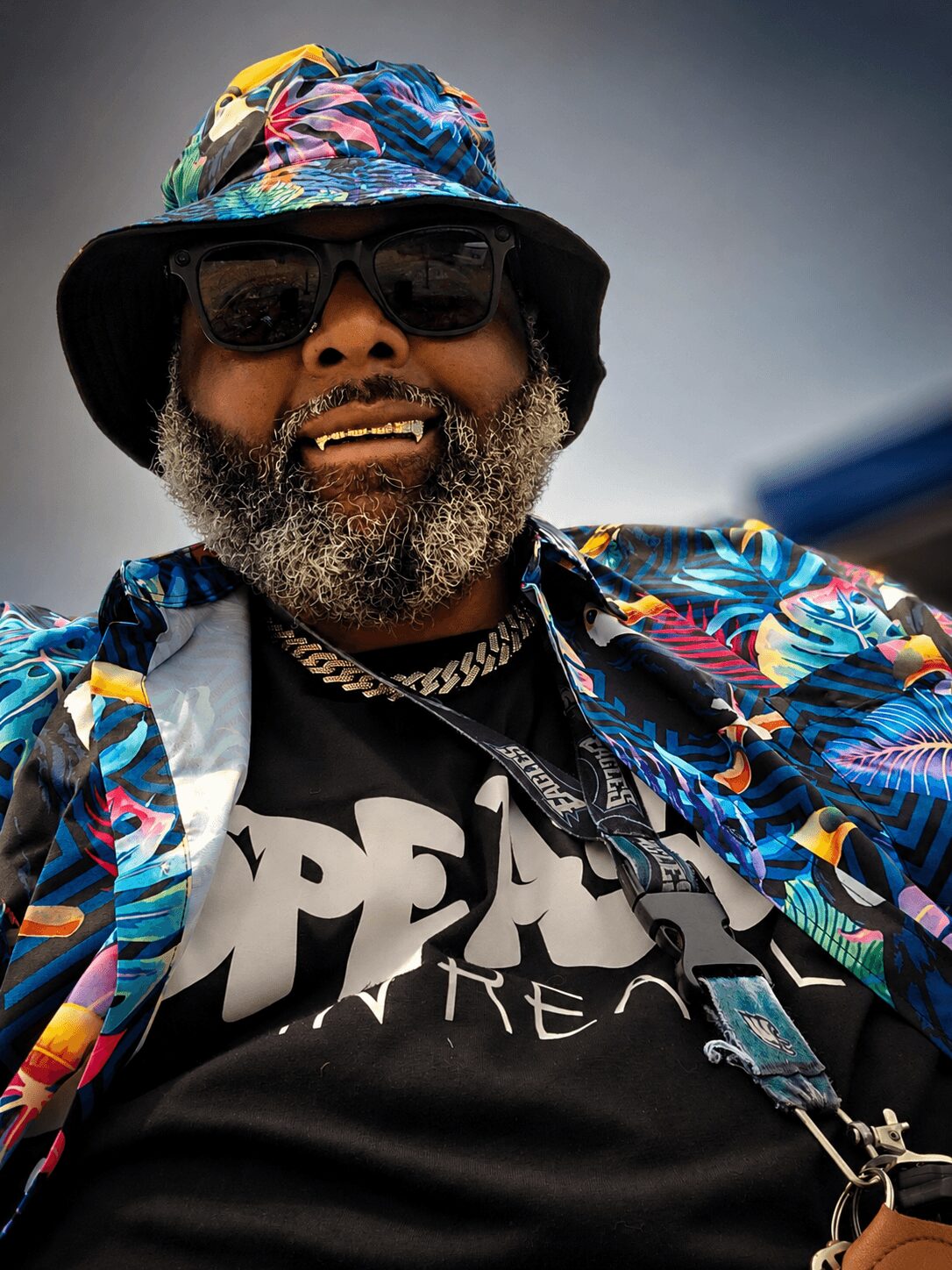 Man with sunglasses, beard, colorful hat, and tropical shirt smiling outdoors.