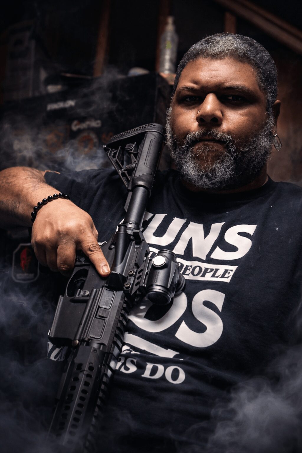 Man with a beard holding a firearm, wearing a black T-shirt with white text, in a dark environment with smoke.