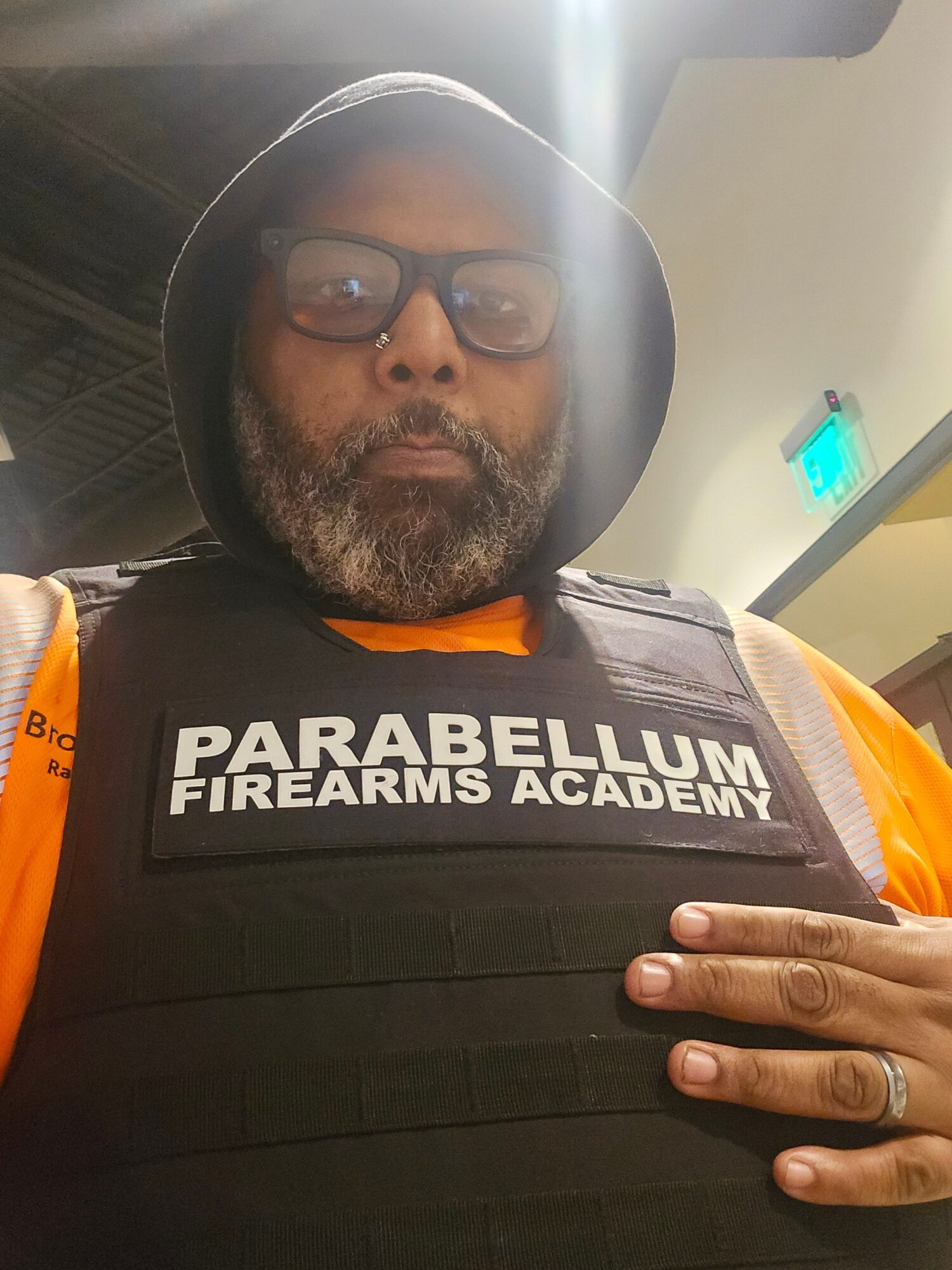 Person wearing a black vest with 'Parabelum Firearms Academy' logo, glasses, and a helmet, indoors with a light source overhead.