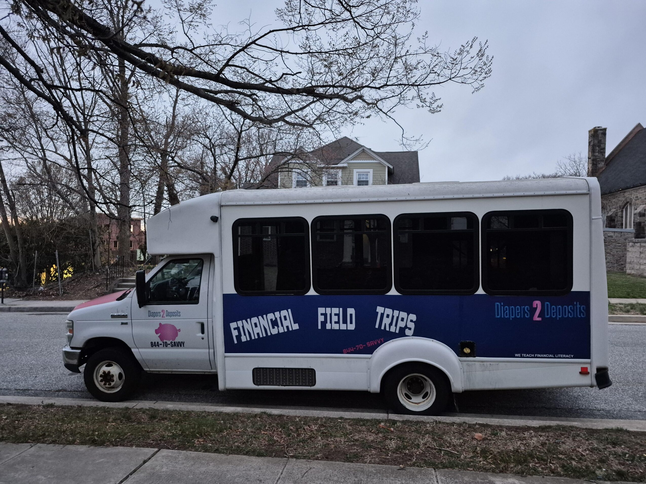 White bus with blue stripe and text, parked on street with houses and trees in background.