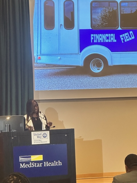 Person speaking at podium with MedStar Health logo, large screen behind showing a blue vehicle with 'Financial Field' text, audience in foreground.