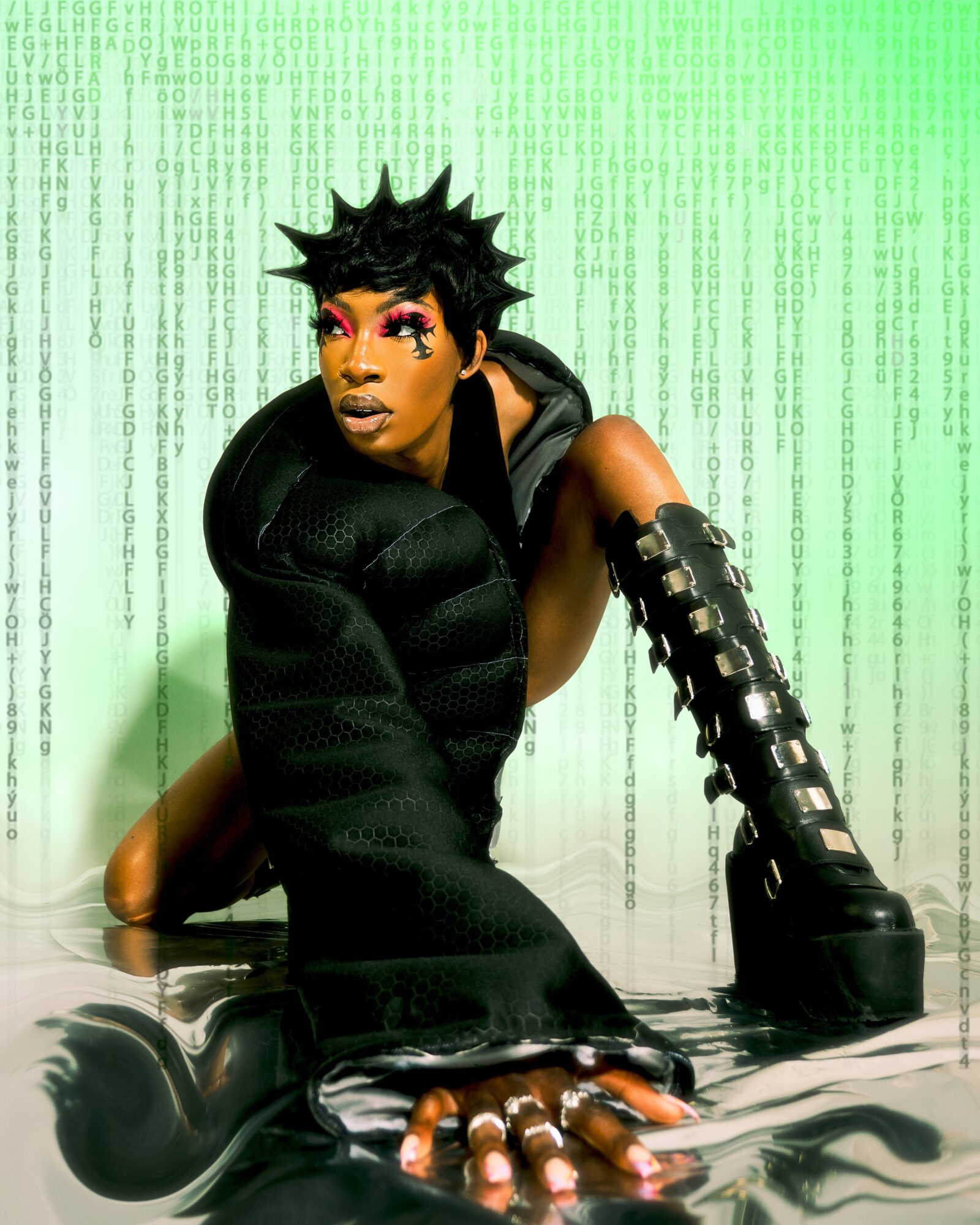 Person with spiky hair, makeup, and futuristic clothing poses against a green digital background, leaning forward with one hand on the floor.