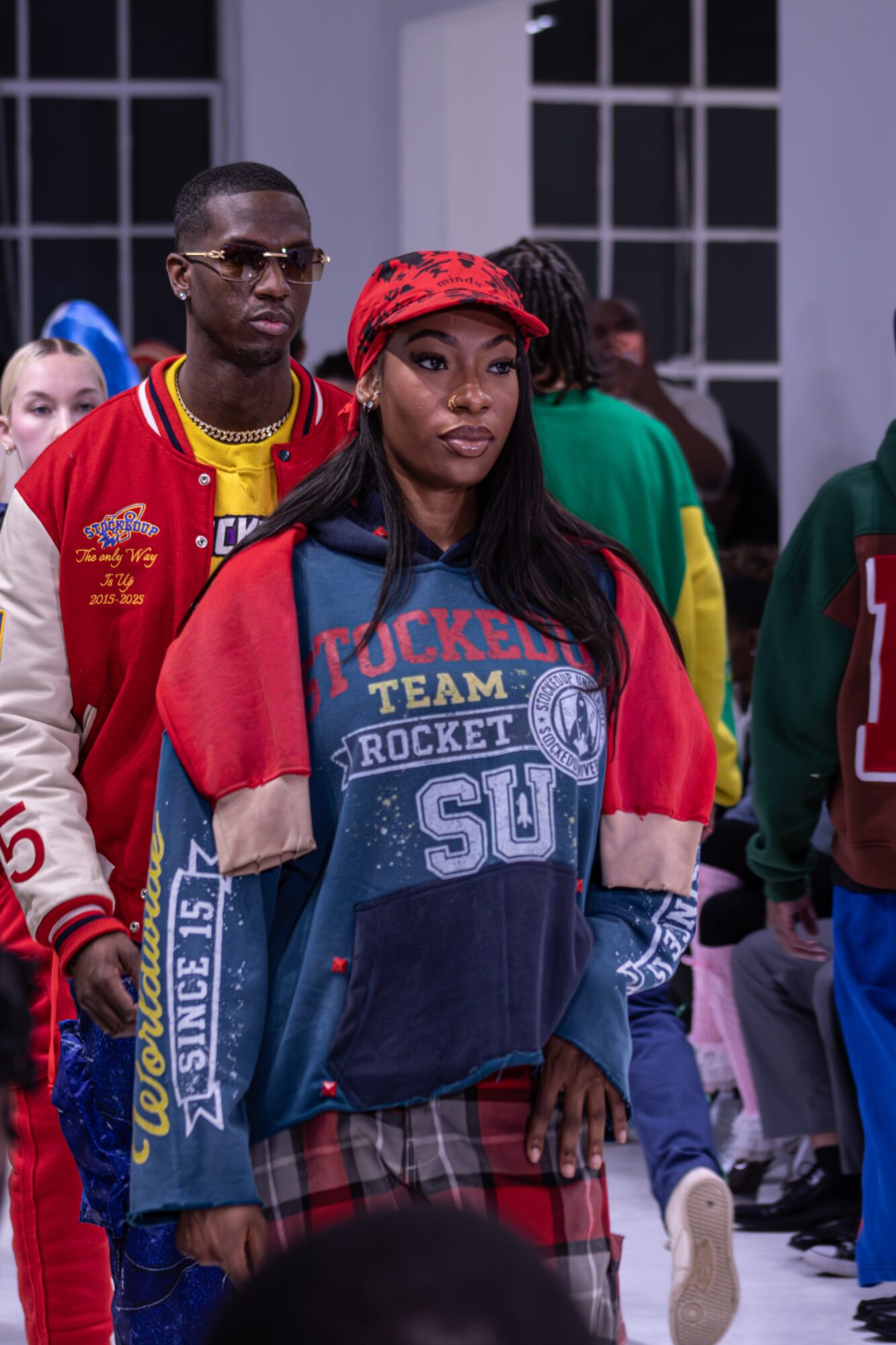 Group of young people indoors, woman in front wearing a red headscarf and colorful jacket, man behind her in sunglasses and red jacket.
