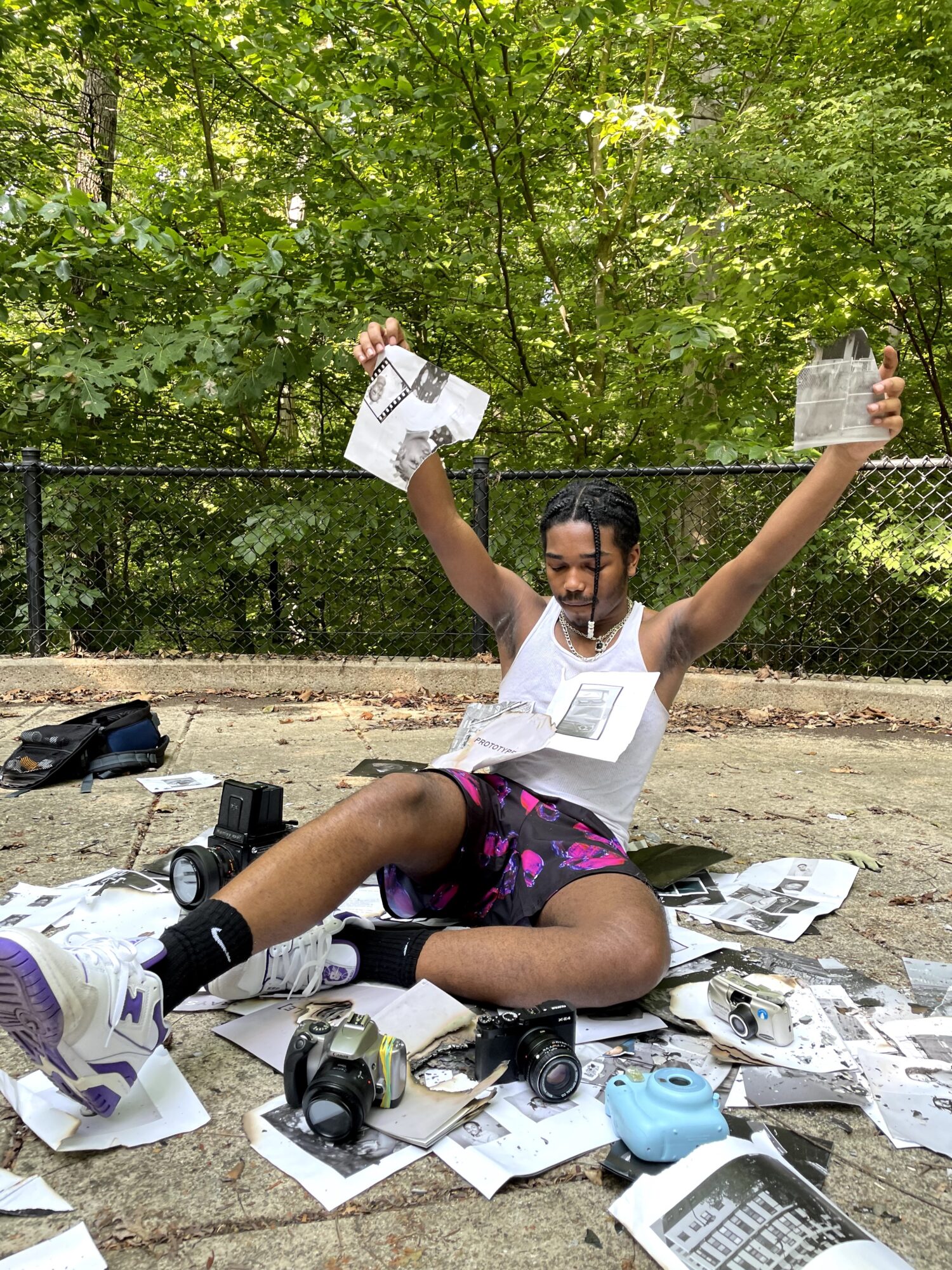 Person sitting on ground outdoors with arms raised, surrounded by scattered papers and cameras, trees in background.