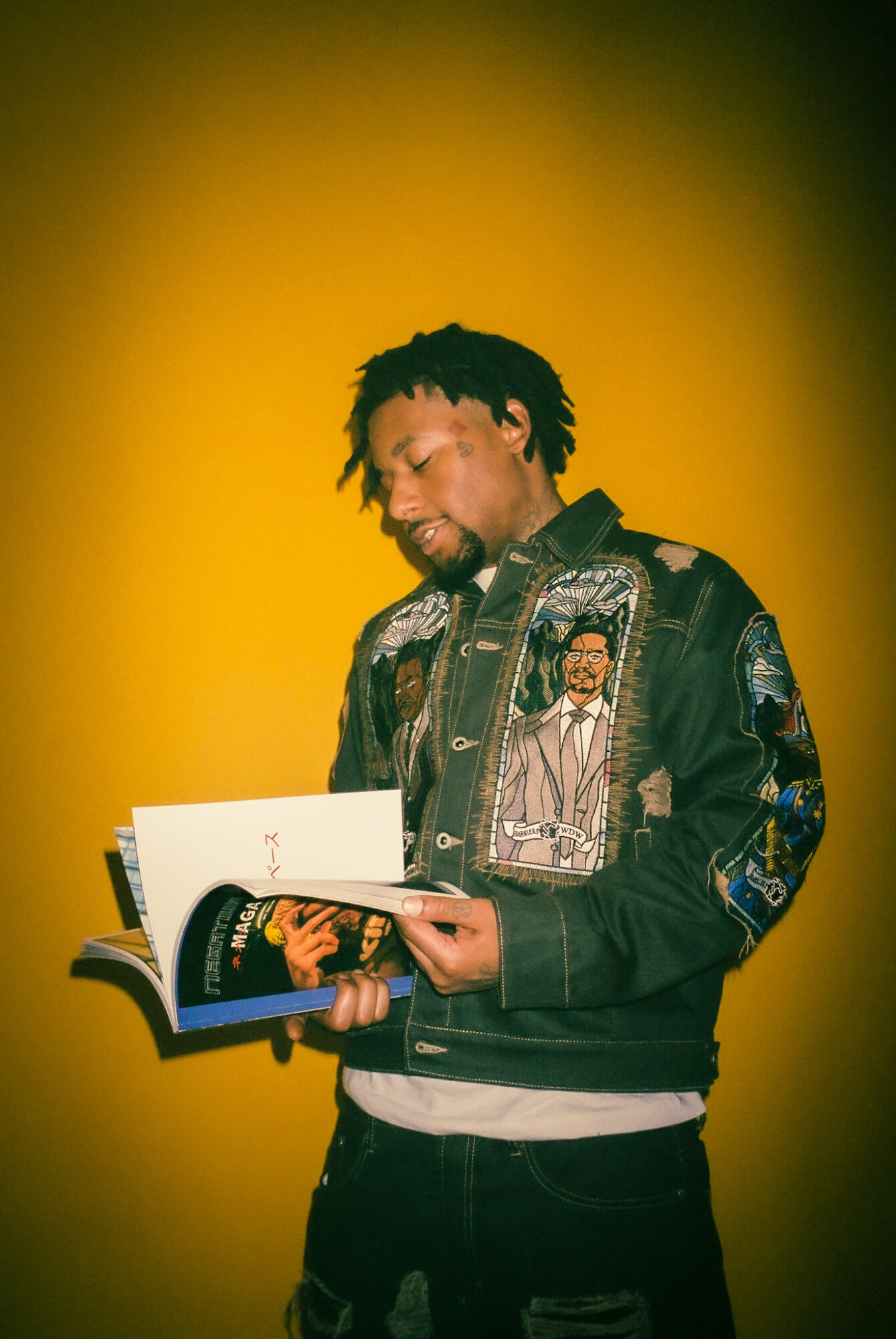 Young man with dreadlocks reading a book, wearing a jacket with patches, against a yellow background.