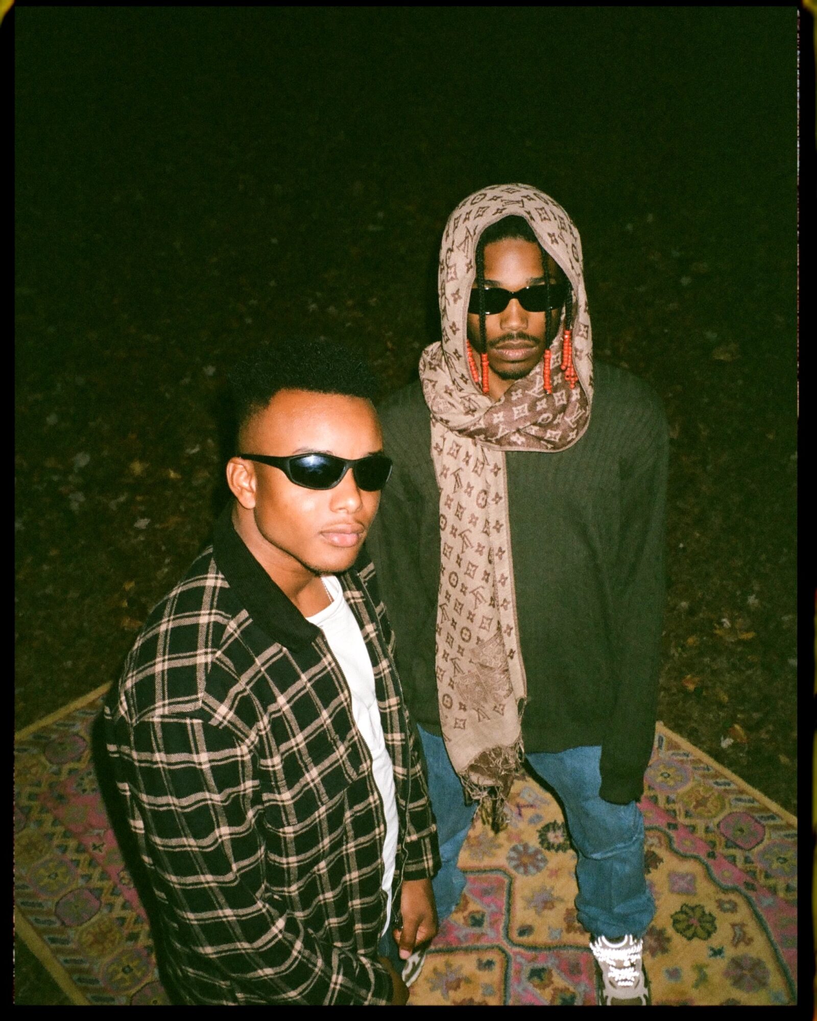 Two people wearing sunglasses stand on a patterned rug outdoors at night, with a dark background.