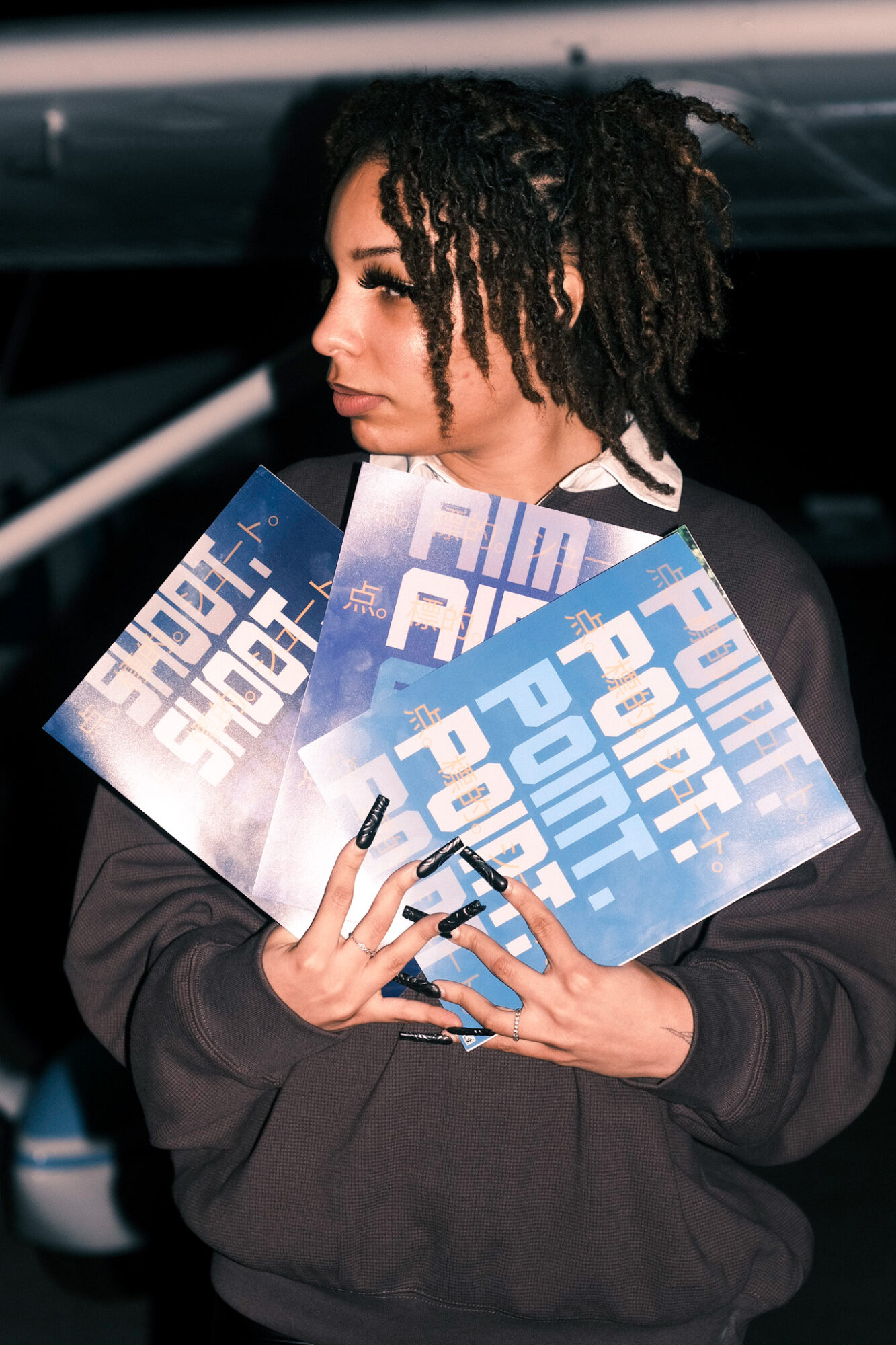 Person with dreadlocks holding flyers, standing outdoors at night, looking to the side, dark background.