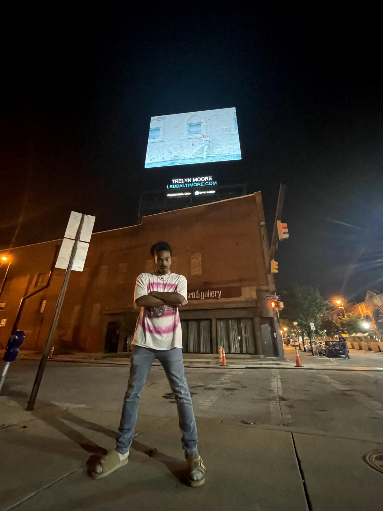 Person standing on sidewalk at night with arms crossed, in front of a building and illuminated billboard, streetlights visible.