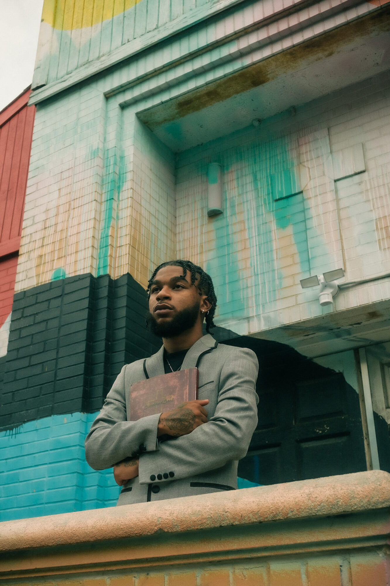 Young man with braided hair in a gray blazer holding a book, standing outdoors near a colorful building.