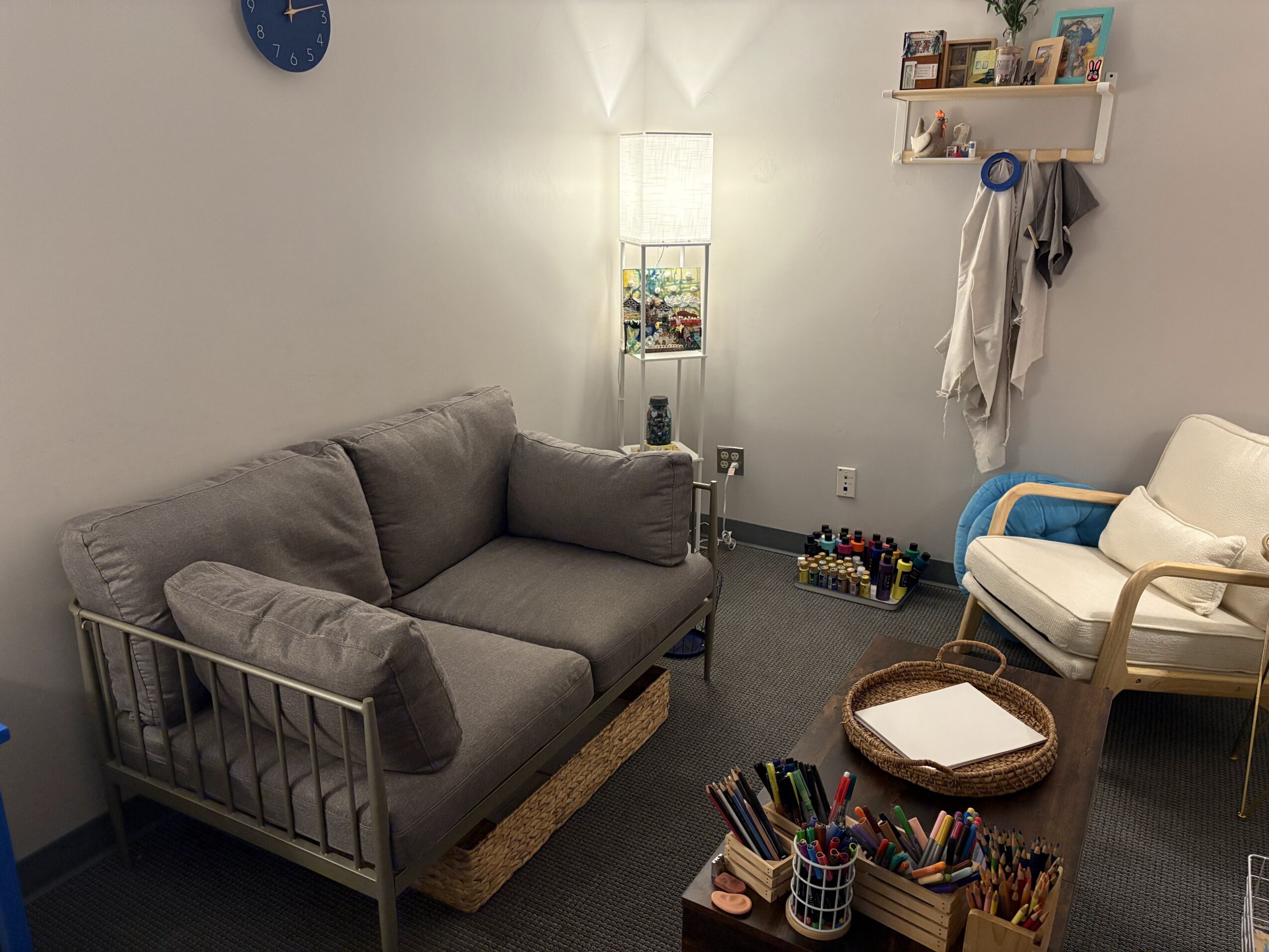 Living room with gray sofa, white armchair, wall shelves, and a tall lamp, with various items on shelves and floor.