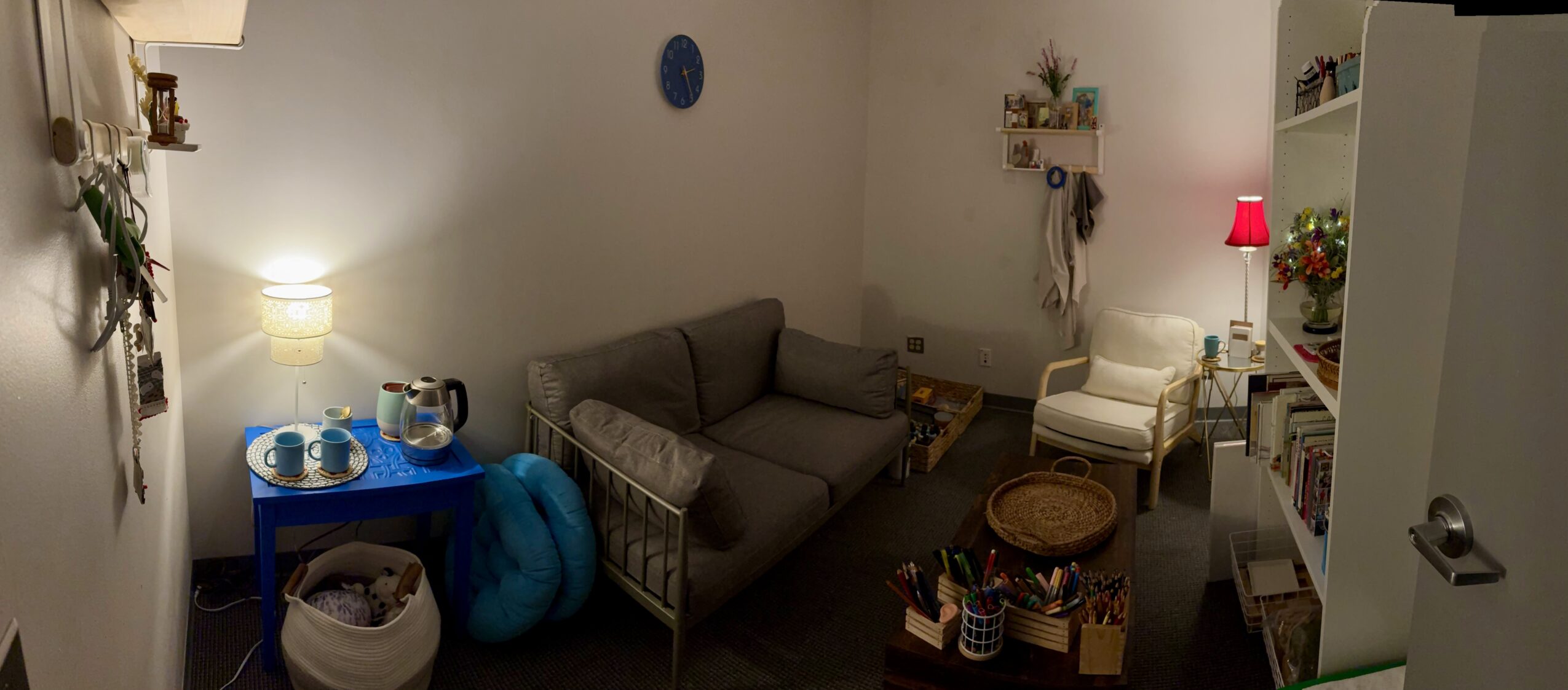 Living room with a gray sofa, white chair, bookshelf, and various decorations, illuminated by lamps and natural light.