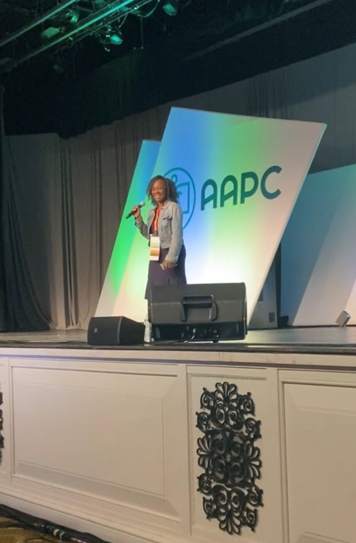 Person standing on stage with a microphone, behind large colorful panels with the APC logo, in a conference setting.