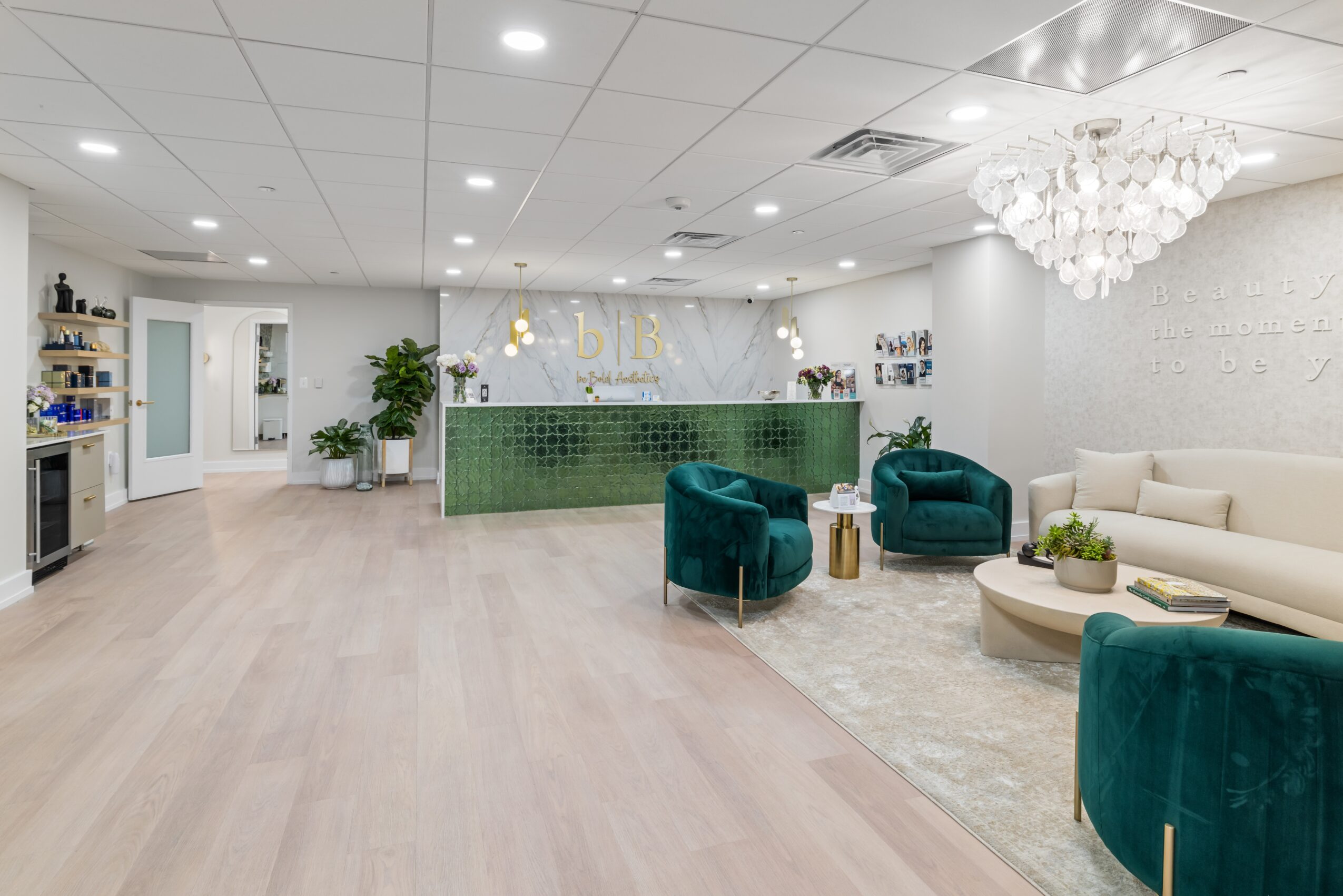 Reception area with a green counter, white walls, teal chairs, a chandelier, and a beige sofa, with light wood flooring.