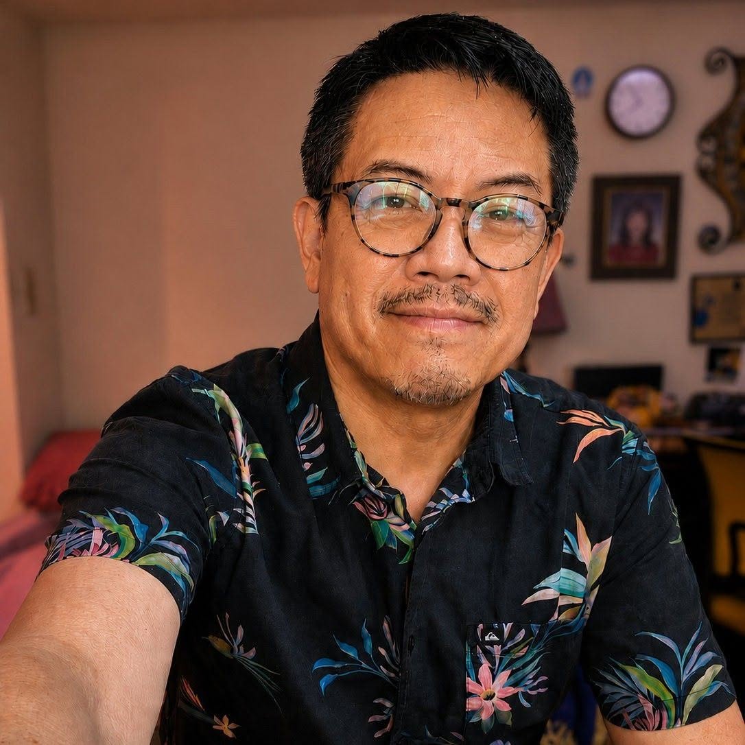 Man with glasses and short dark hair wearing a floral shirt, smiling in a room with clocks and pictures on the wall.
