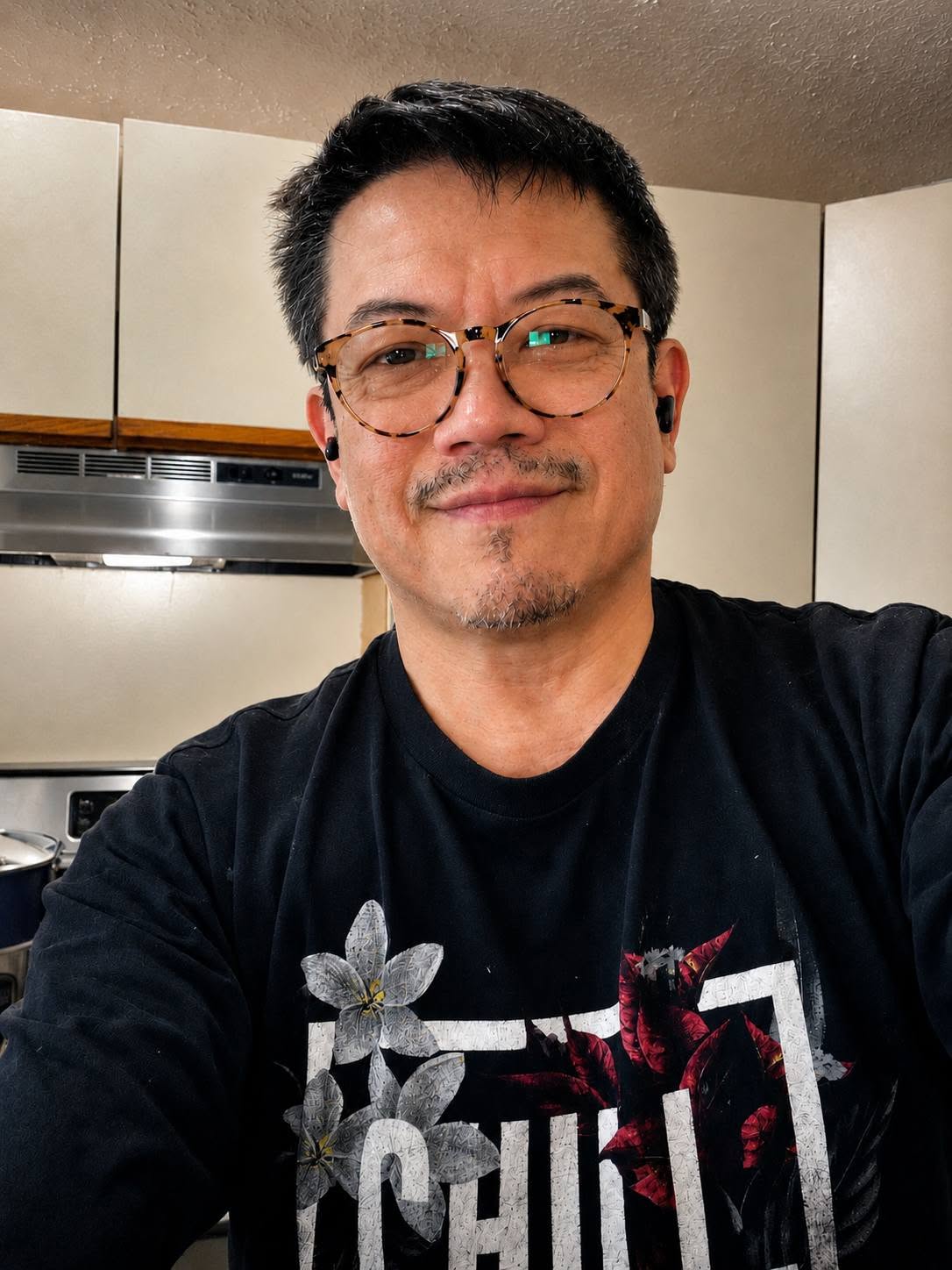 Man with glasses and short dark hair smiling indoors, kitchen background, wearing a black T-shirt with a floral design.