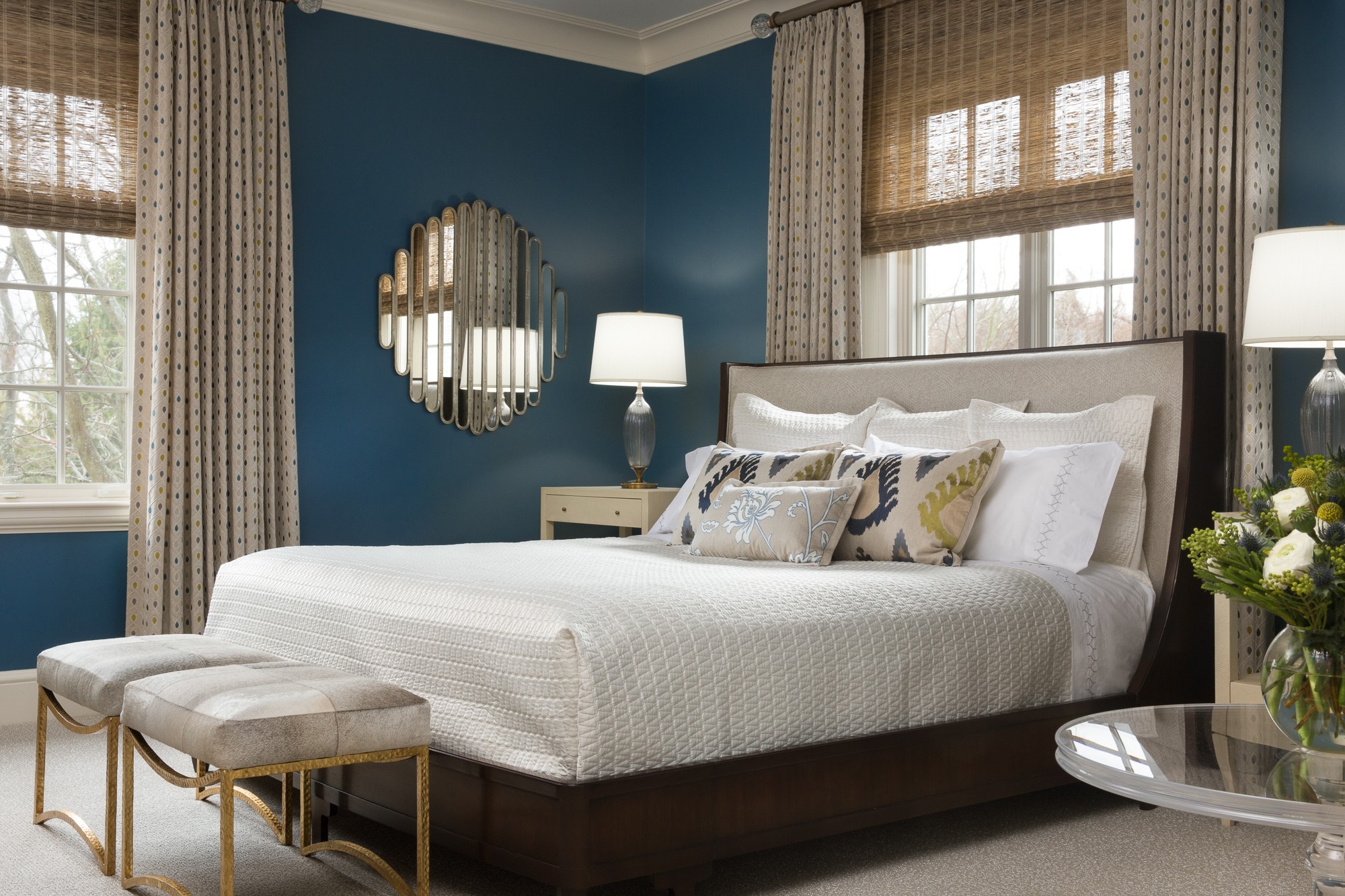 Bedroom with blue walls, white bed, two lamps, window with curtains, and a round mirror, decorated in neutral tones.