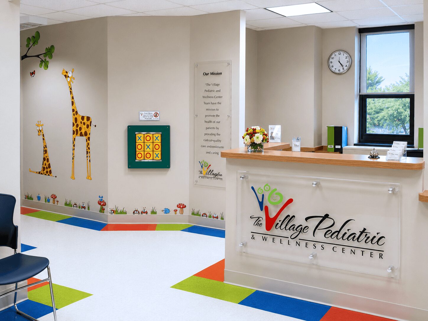 Reception area with giraffe wall decals, colorful floor tiles, a clock, and a window, in a pediatric wellness center.