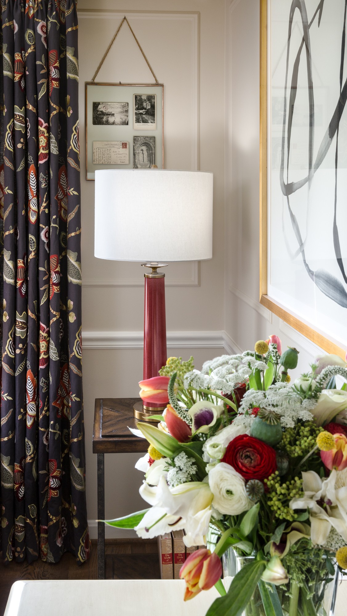 A bouquet of flowers in front of a window with a large abstract painting, a table lamp, and a framed picture on the wall.
