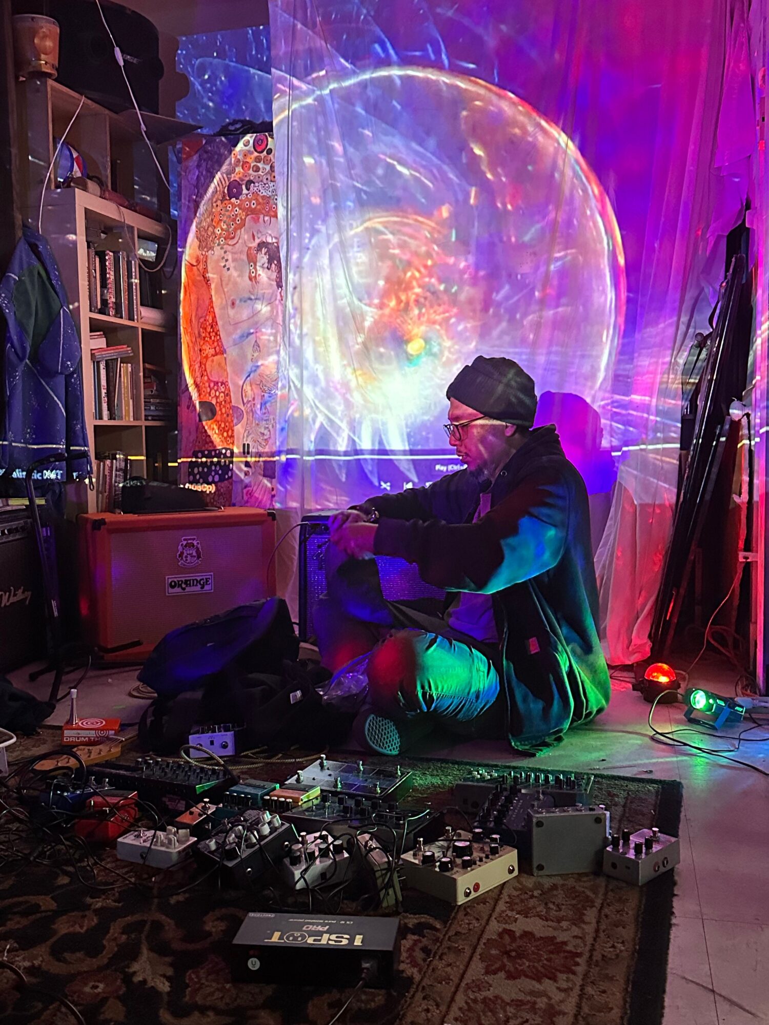 Person crouching near DJ equipment with colorful lights and a large circular projection in the background.