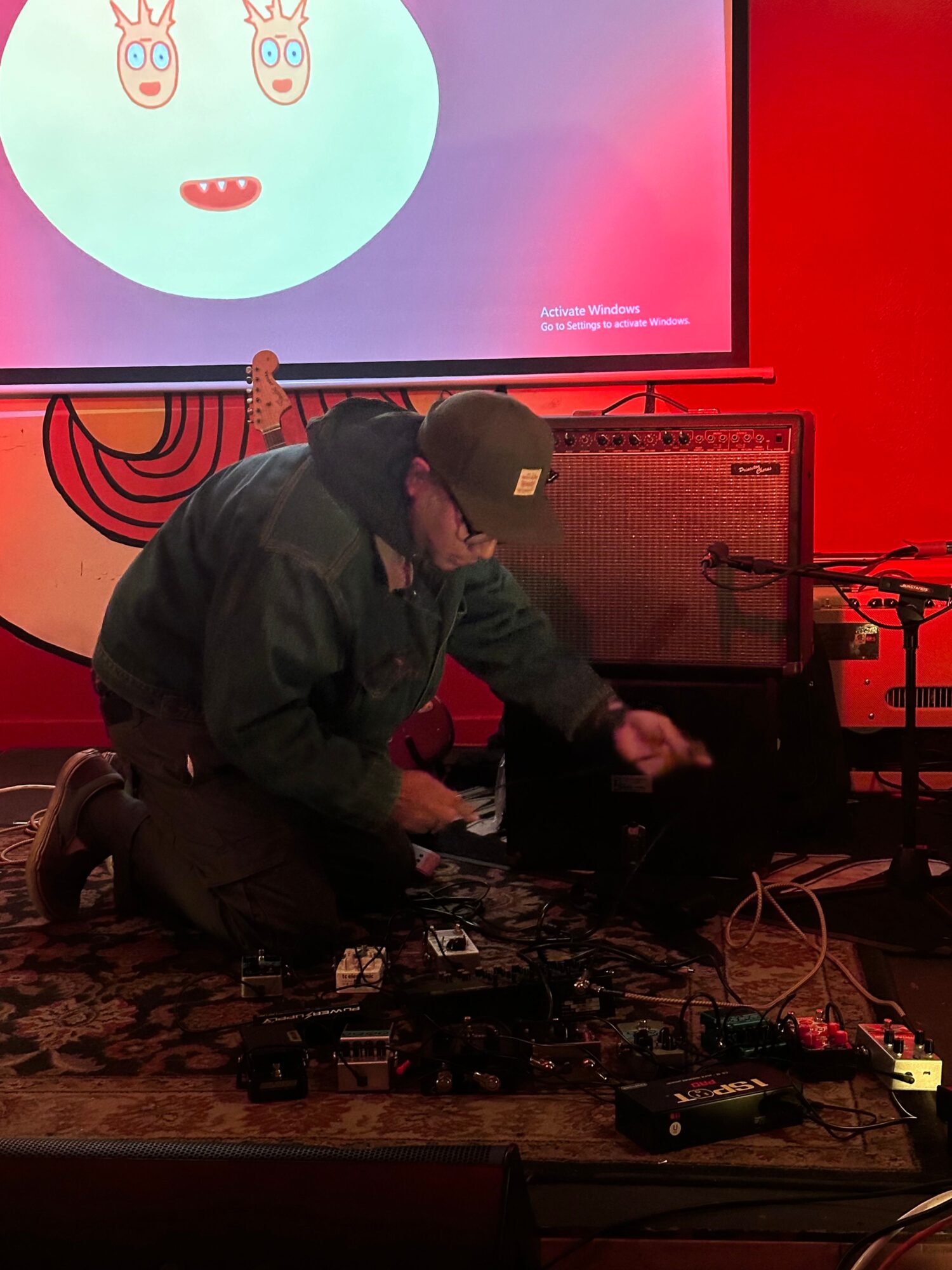 Person sitting on the floor with a microphone, surrounded by electronic equipment, in a room with a red wall and screen.