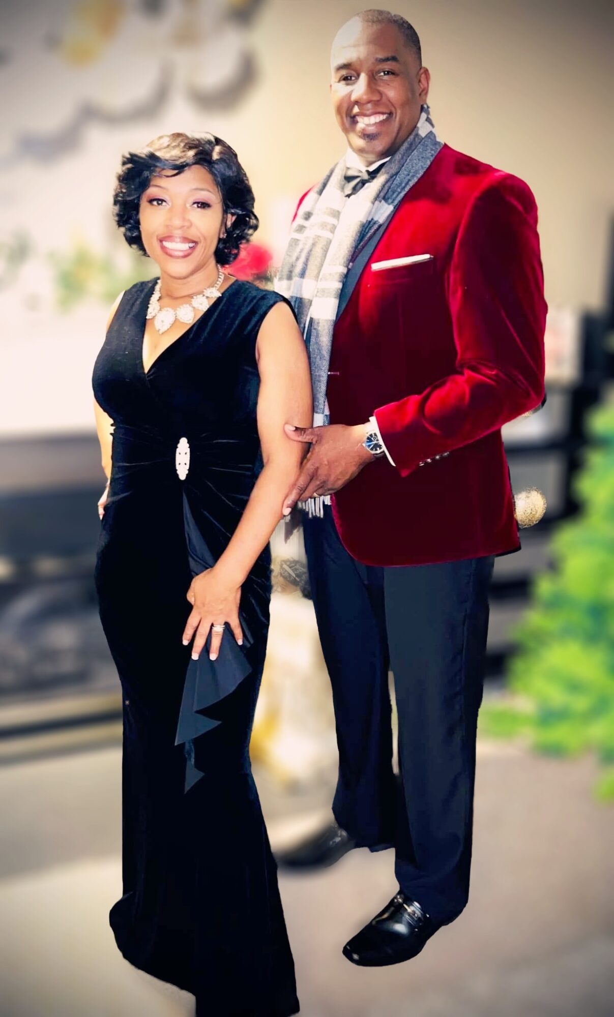 A smiling man and woman stand together indoors, dressed formally, with the woman in a black dress and the man in a red blazer.