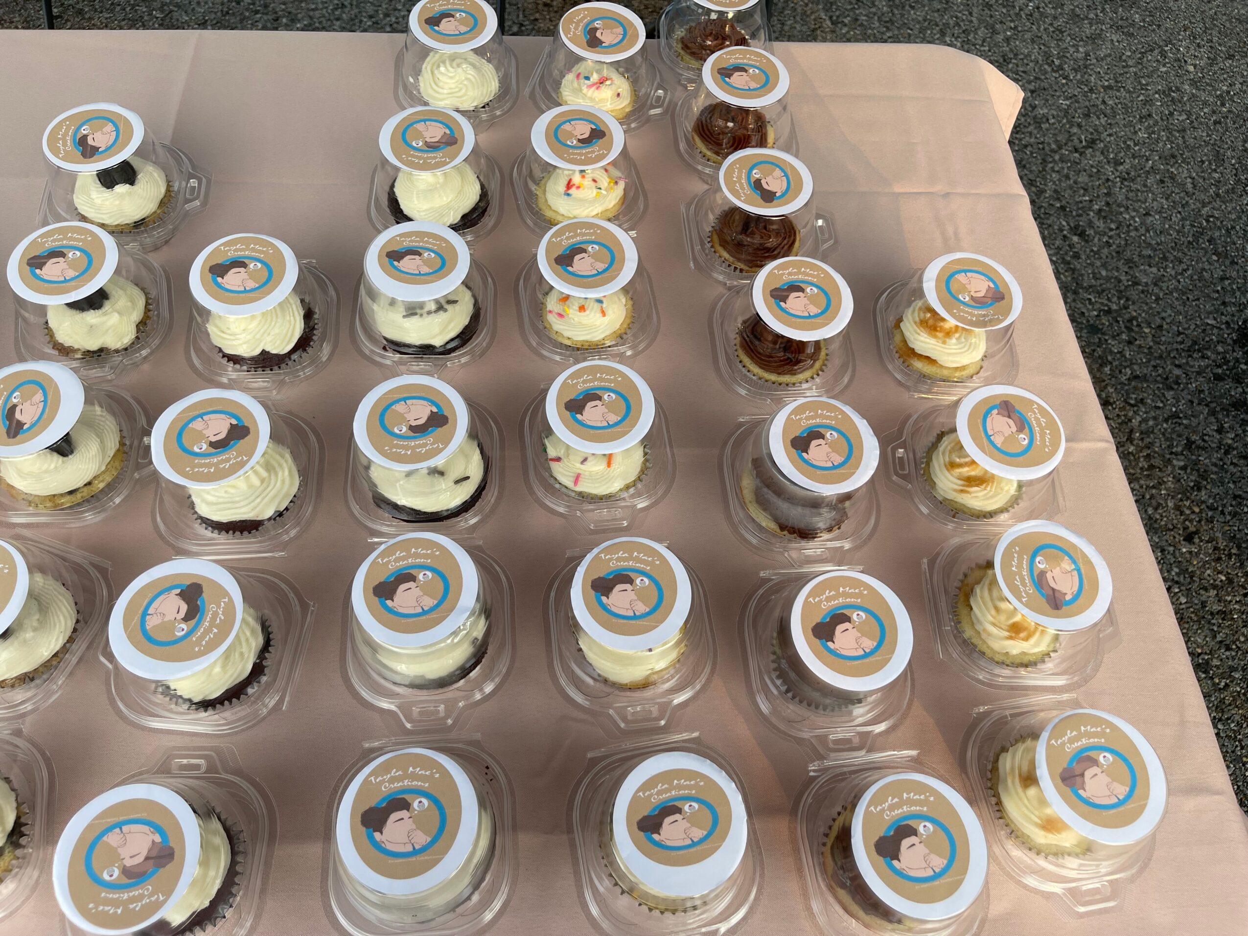 Rows of cupcakes with white frosting and circular toppers on a table, some with brown topping, on a pink tablecloth.