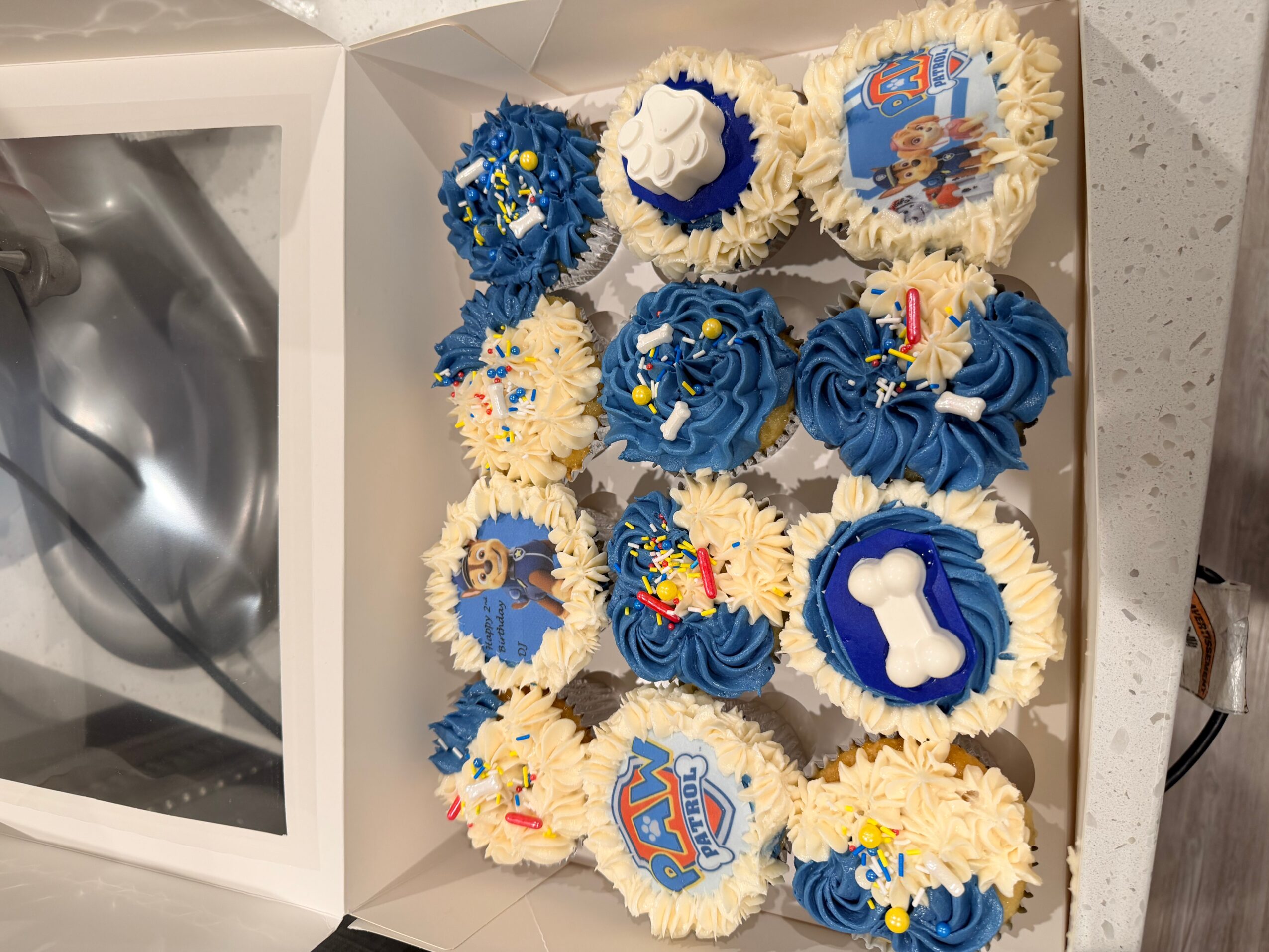 Box of decorated cupcakes with blue and white themes, some with character images and dog bone shapes, on a table.