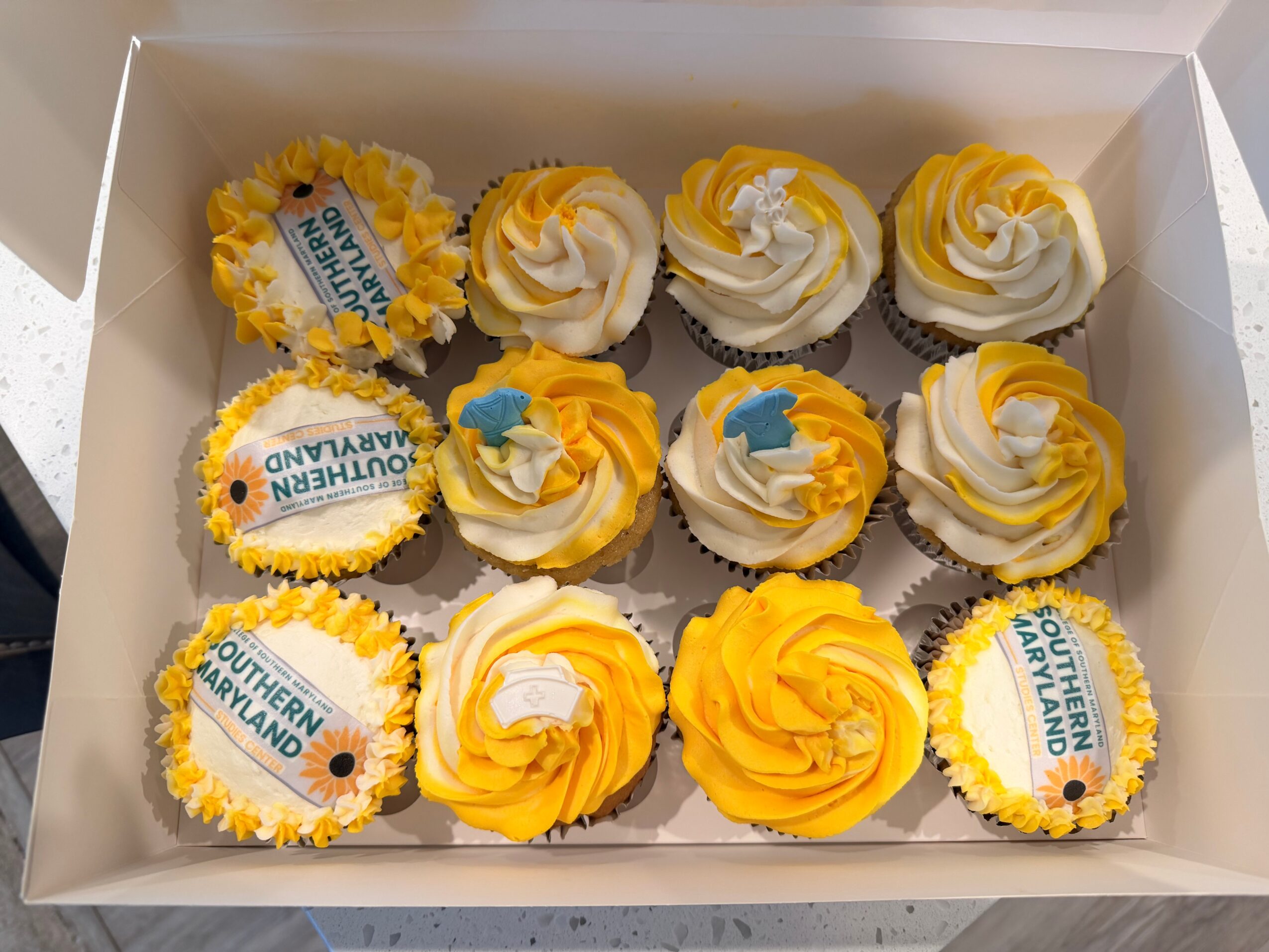 Box of twelve cupcakes with yellow and white frosting, some decorated with blue and white edible toppers, arranged in three rows.