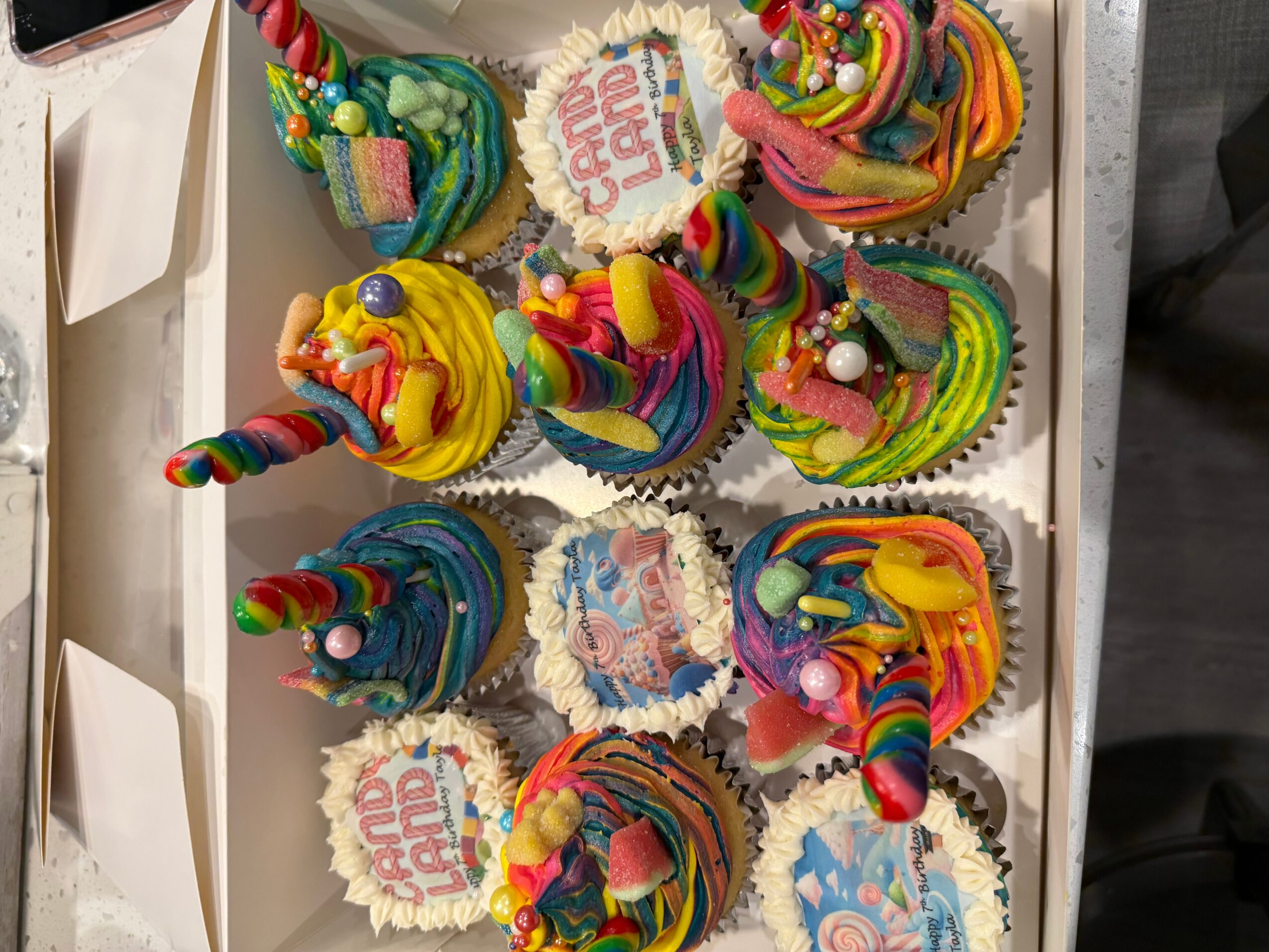 A tray of colorful cupcakes with rainbow frosting, sprinkles, and candles, some with birthday messages on toppers.