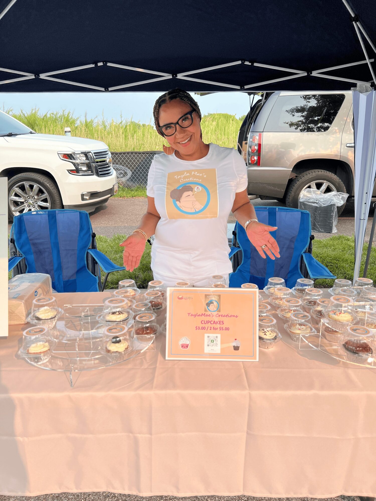 Young woman with glasses standing behind a table with baked goods under a canopy, cars parked behind her.