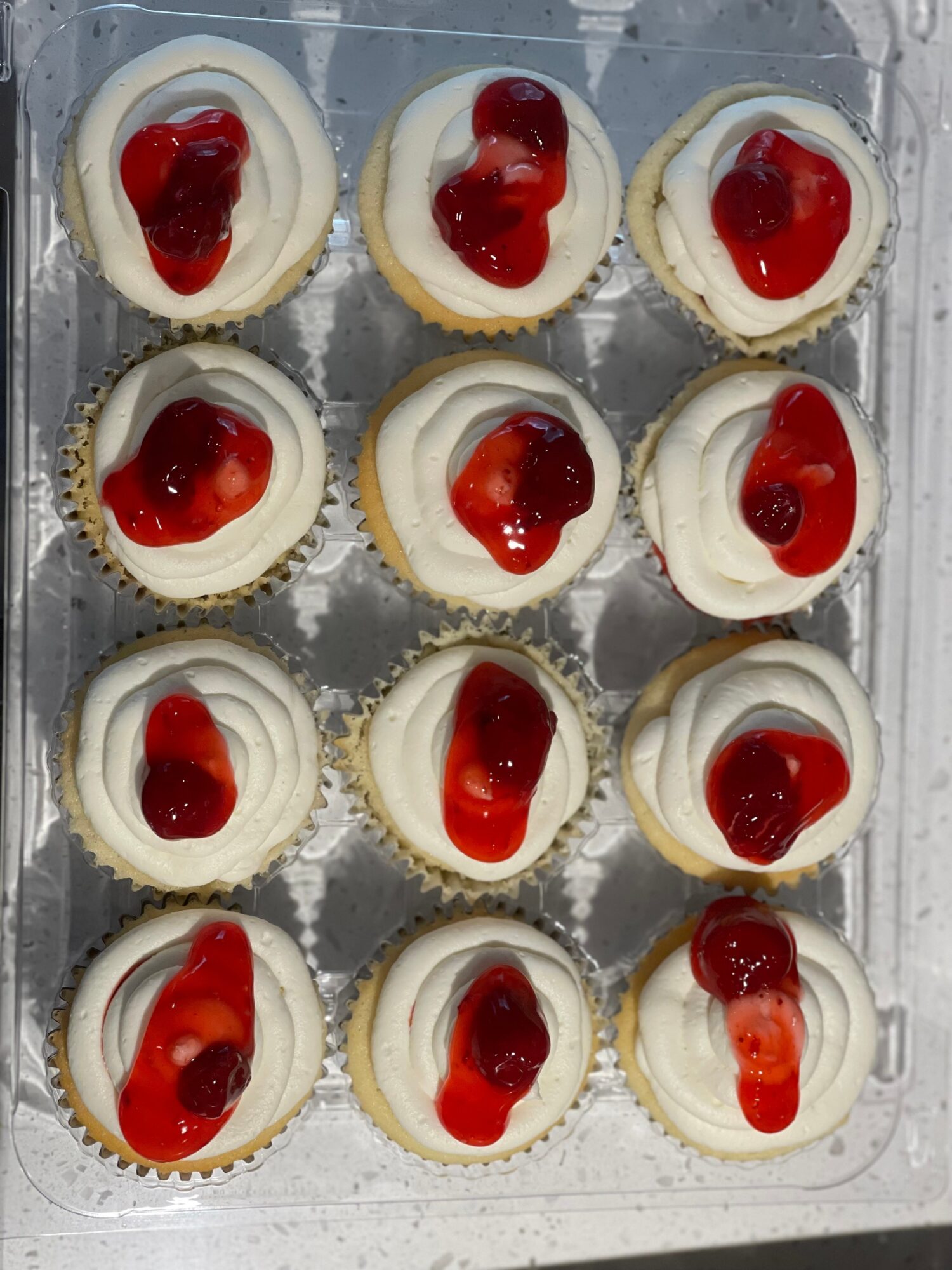 Twelve cupcakes with white frosting and red cherry toppings arranged in a 3x4 grid on a clear tray.
