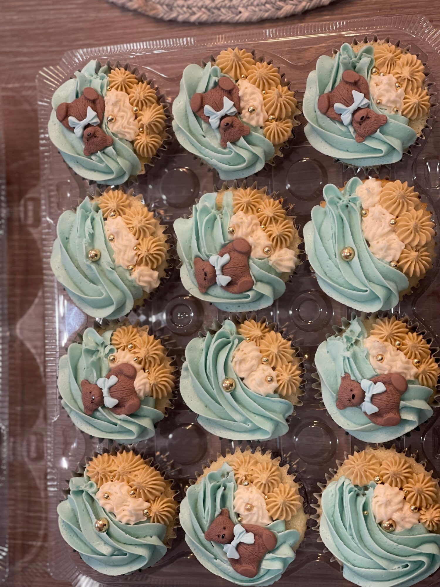 Tray of decorated cupcakes with teddy bear and swirl frosting, in pastel colors, on a clear plastic tray.