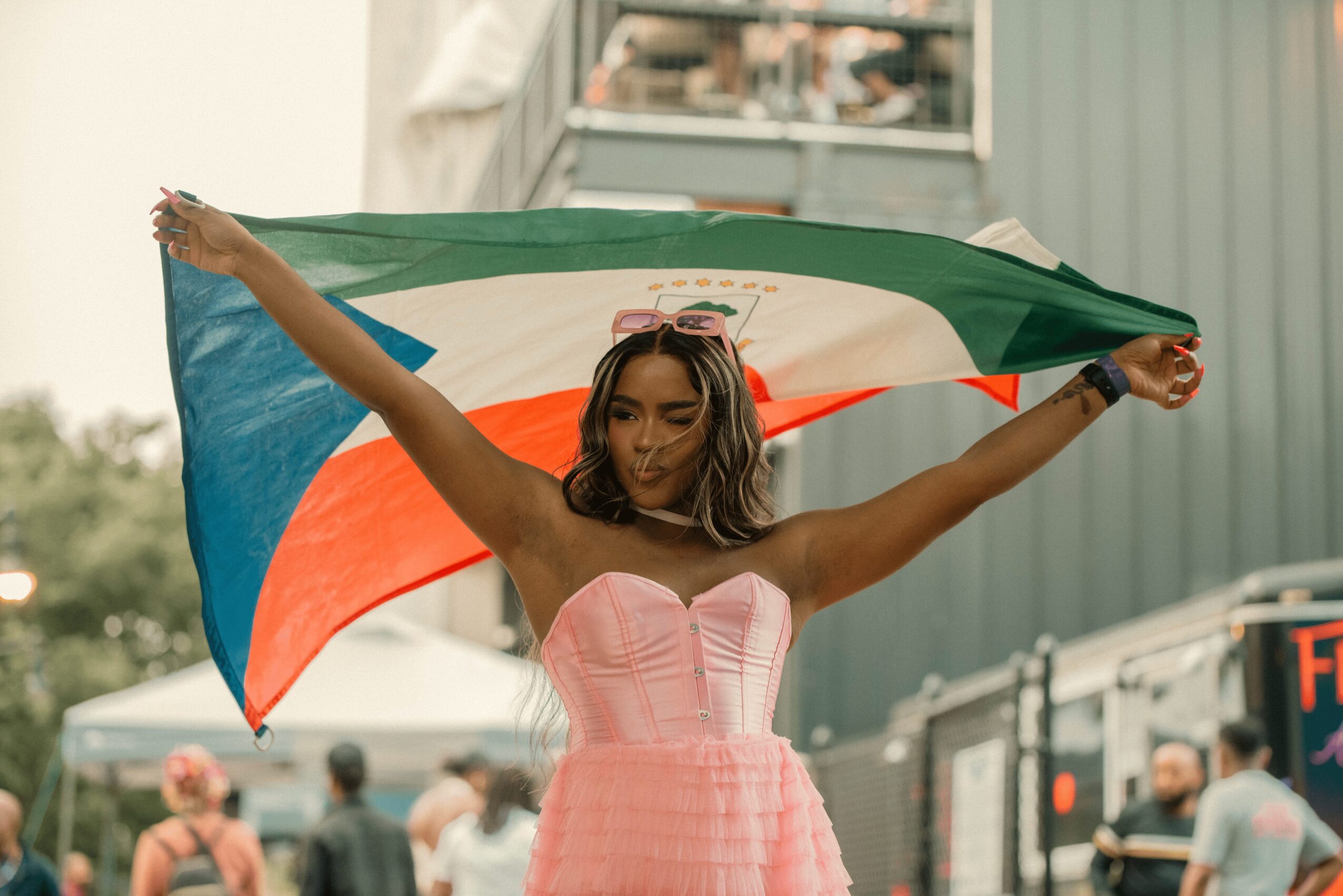 Woman holding an Italian flag outdoors, wearing a pink strapless dress, with a crowd and vehicles in the background.
