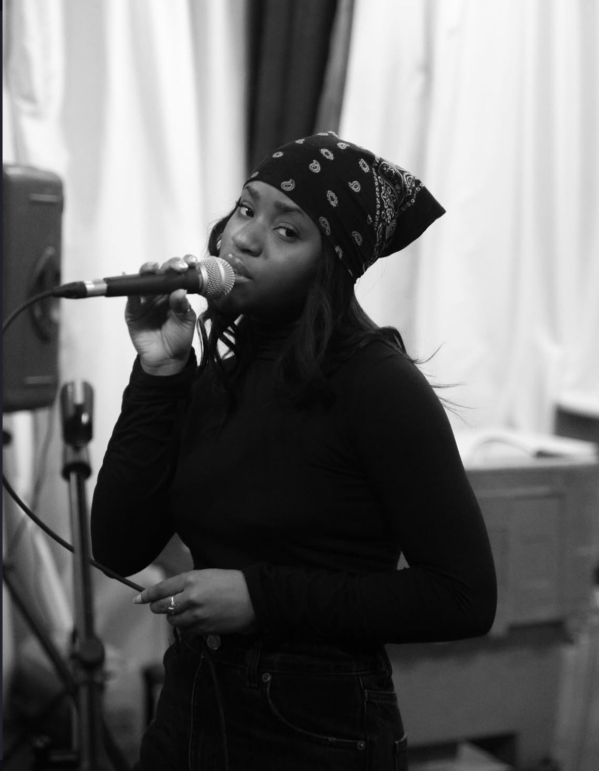 Woman wearing a bandana singing into a microphone in a room with curtains and equipment.