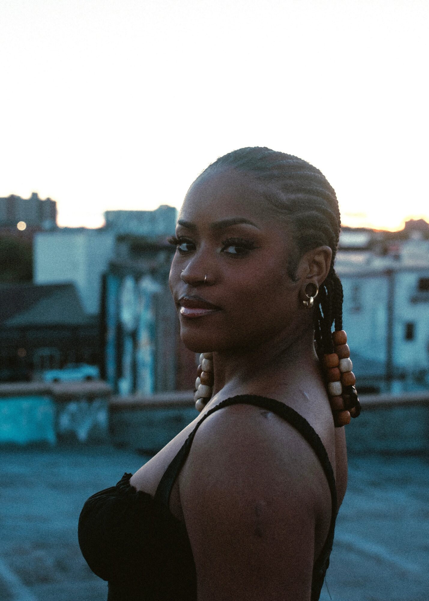 A woman with braided hair and earrings looks over her shoulder against an urban background at sunset.