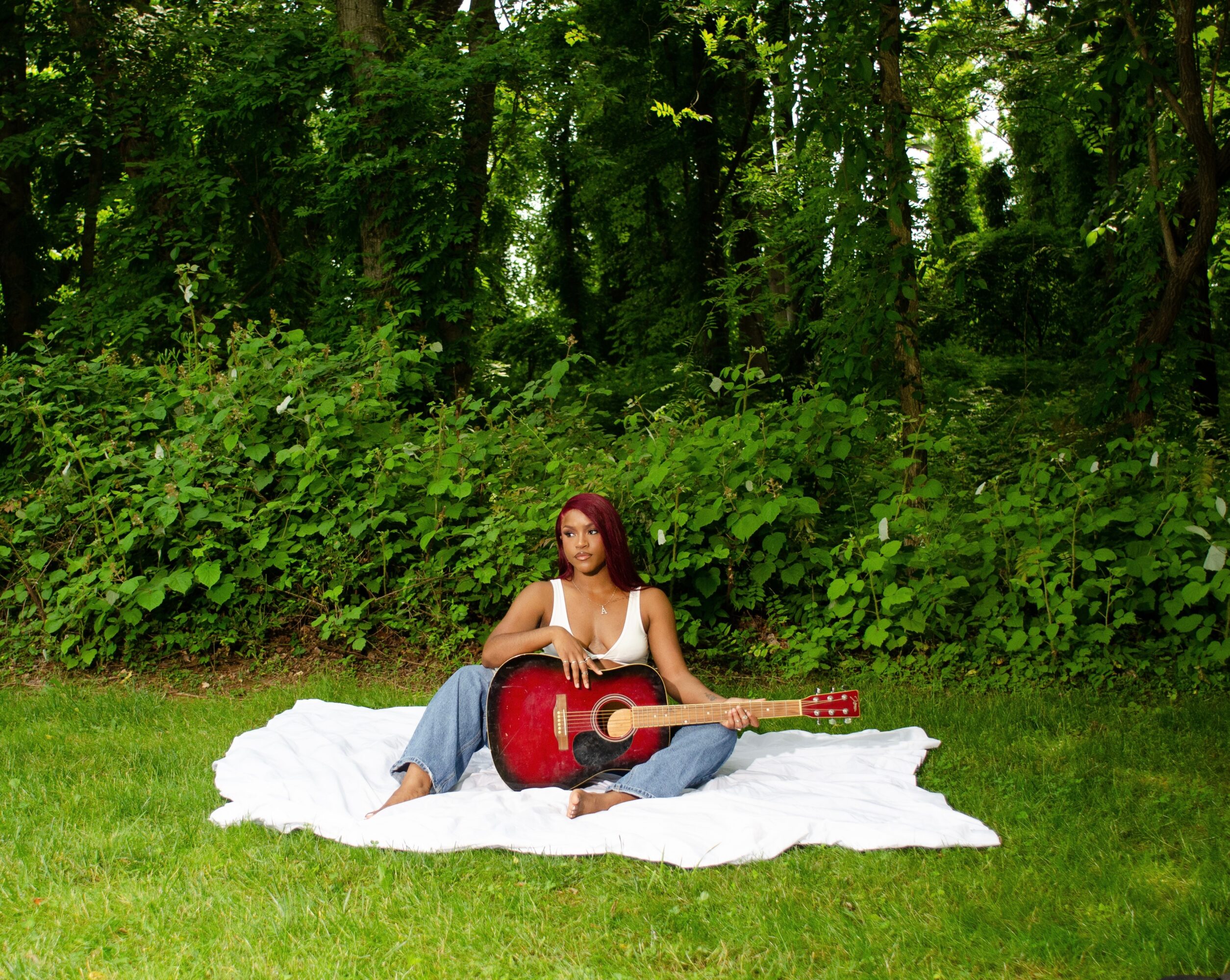 Person sitting on a white blanket playing a red acoustic guitar outdoors with green trees and bushes in the background.