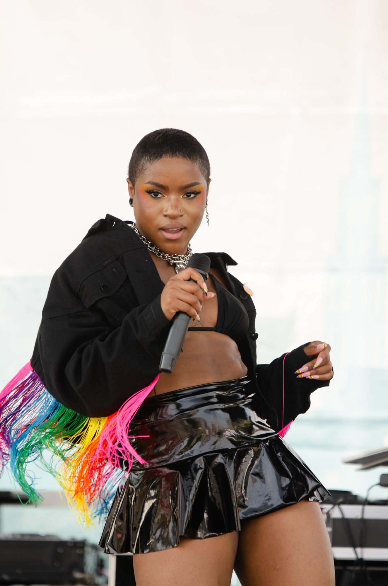 Woman with short hair holding a microphone, wearing a black crop top, shiny black skirt, and a black jacket, with a colorful rainbow hair extension.