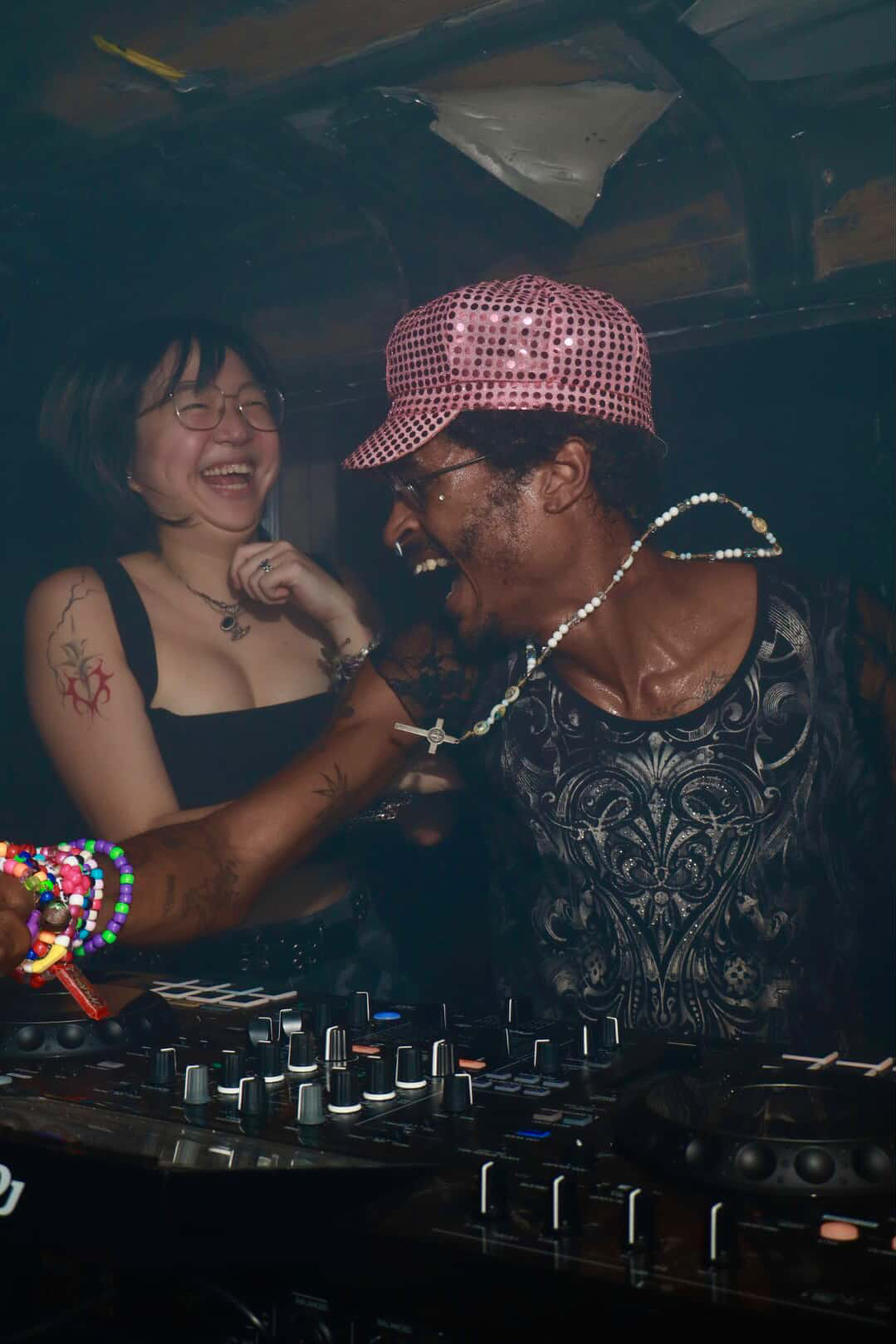 Two people smiling and dancing at a party, one wearing a pink hat and glasses, the other with tattoos and jewelry.
