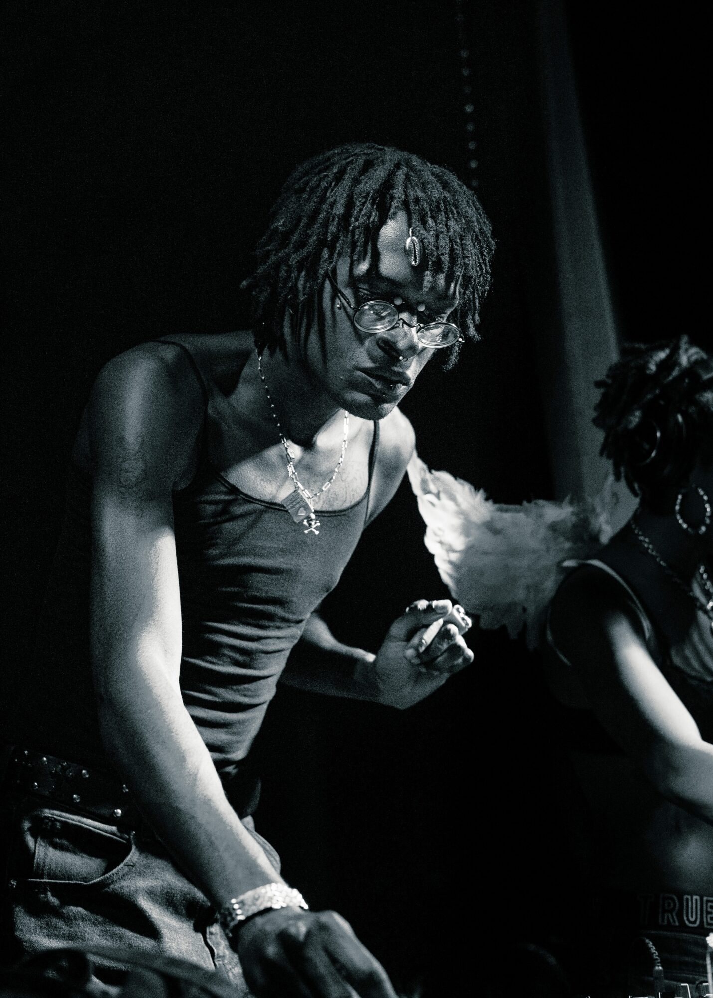 Person with dreadlocks and glasses wearing a tank top, holding a cigarette, in a dark setting.