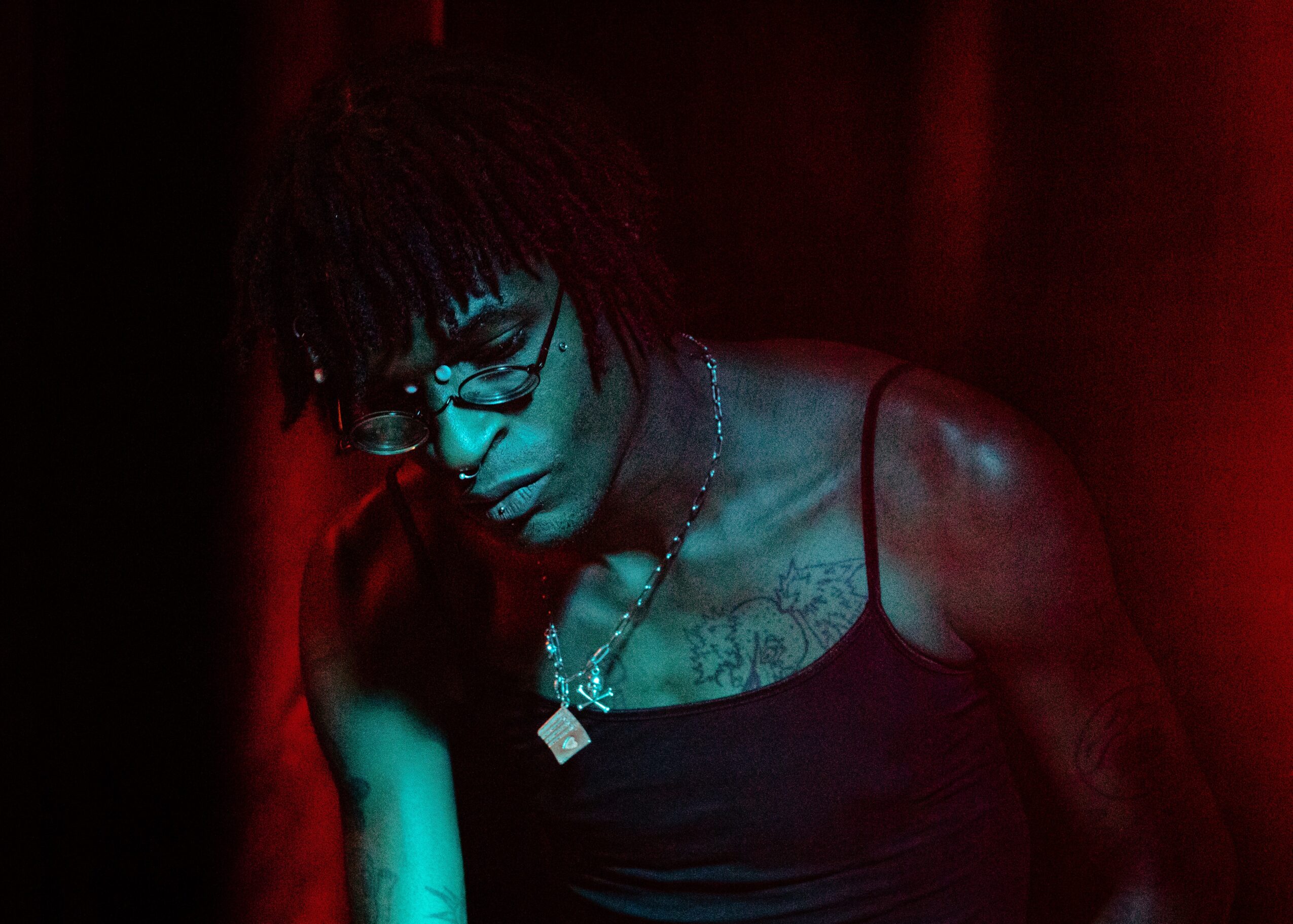Person with glasses and dreadlocks looking down, wearing a black tank top, in a dark red-lit environment.