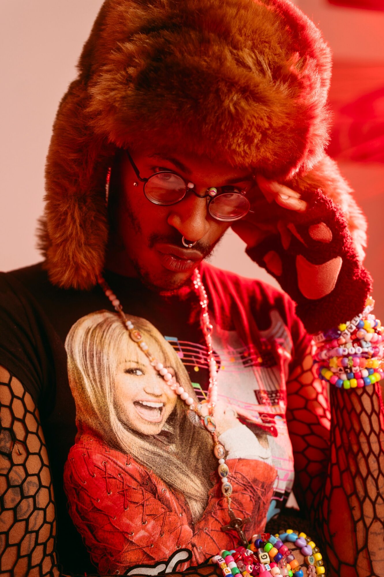 Person with glasses and furry hat adjusting glasses, wearing colorful jewelry and fishnet gloves, with a smiling woman in the background.