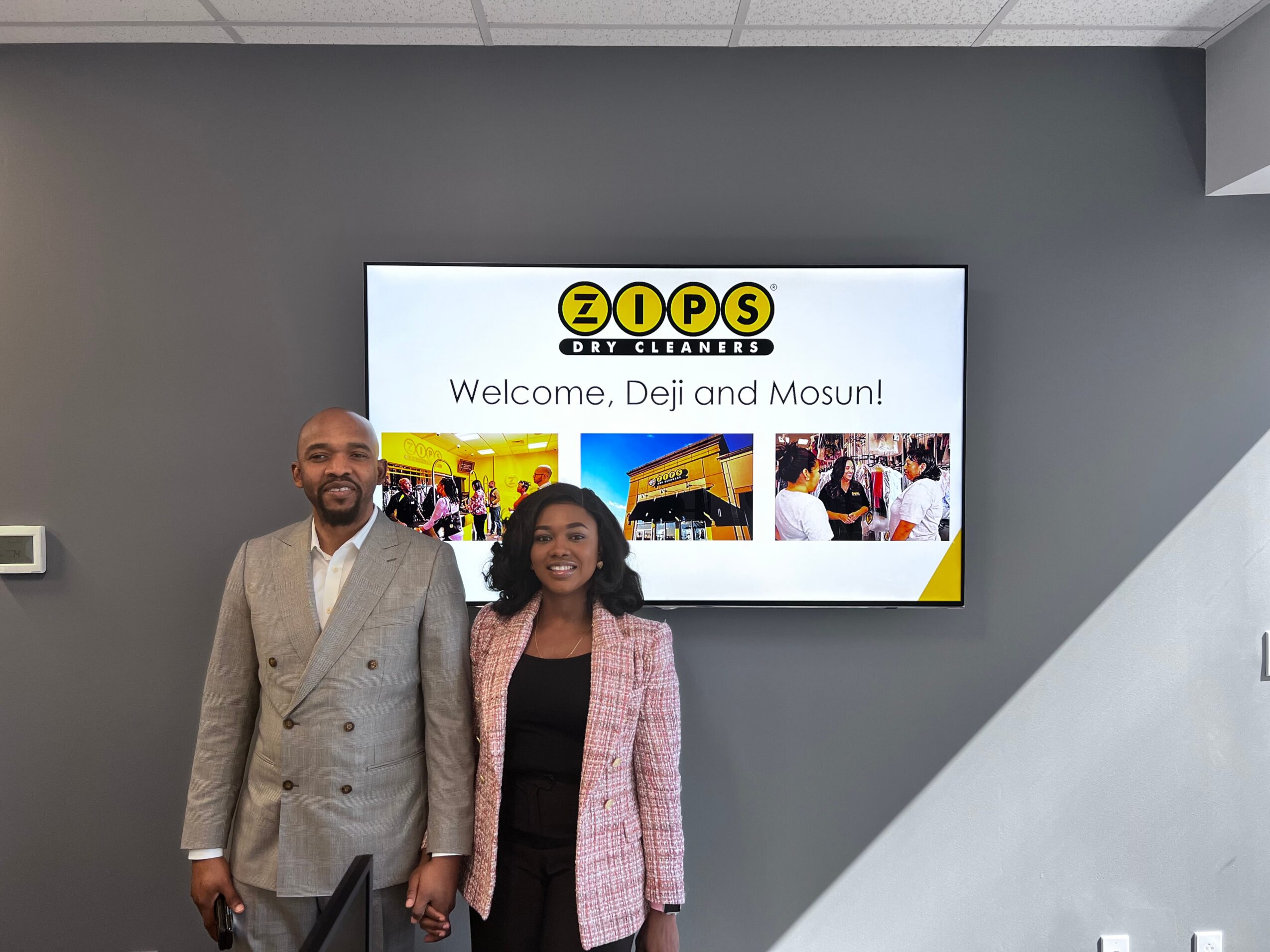 Two people standing in front of a screen displaying a welcome message and photos, holding hands and smiling.