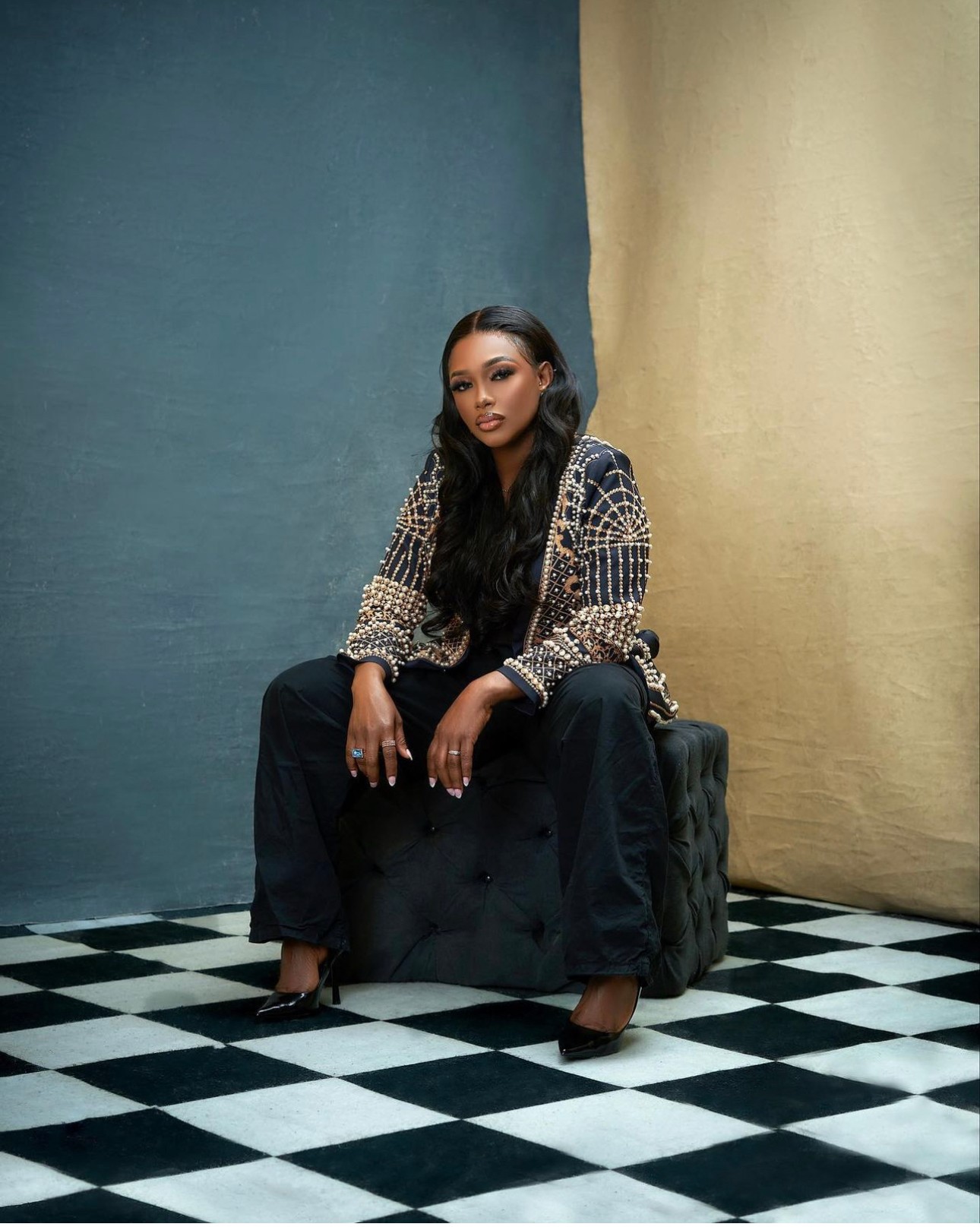 Woman sitting on a black ottoman with a checkered floor and colorful backdrop, wearing a patterned top and dark pants.