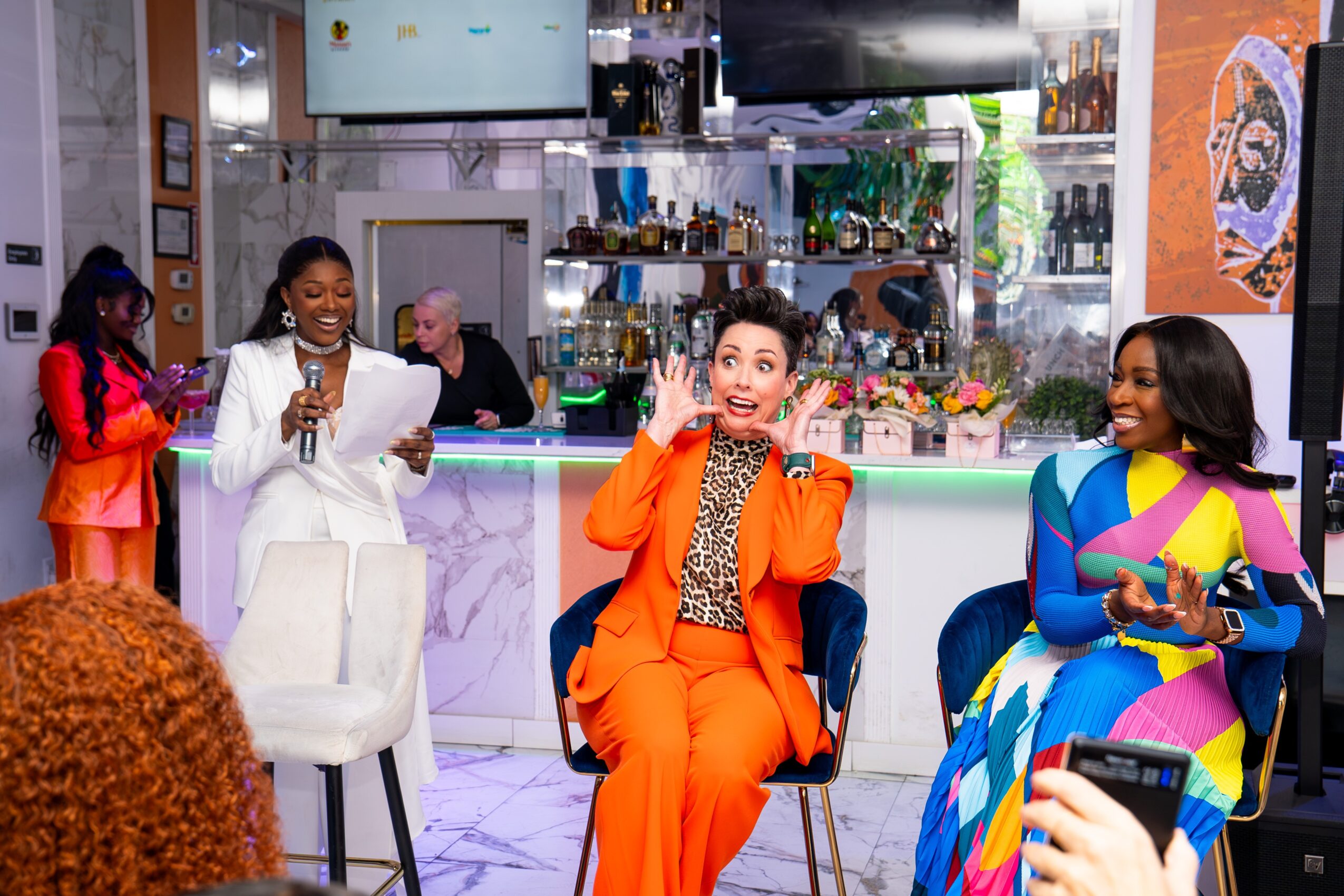 Three women sitting at a bar, smiling and talking, with a woman in a colorful dress on the right, and a woman in an orange suit in the middle, in a lively setting.