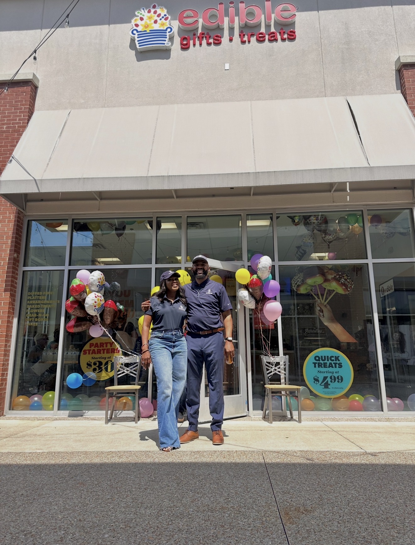 Three people stand outside a store with balloons and chairs, storefront has large glass windows and a sign above.