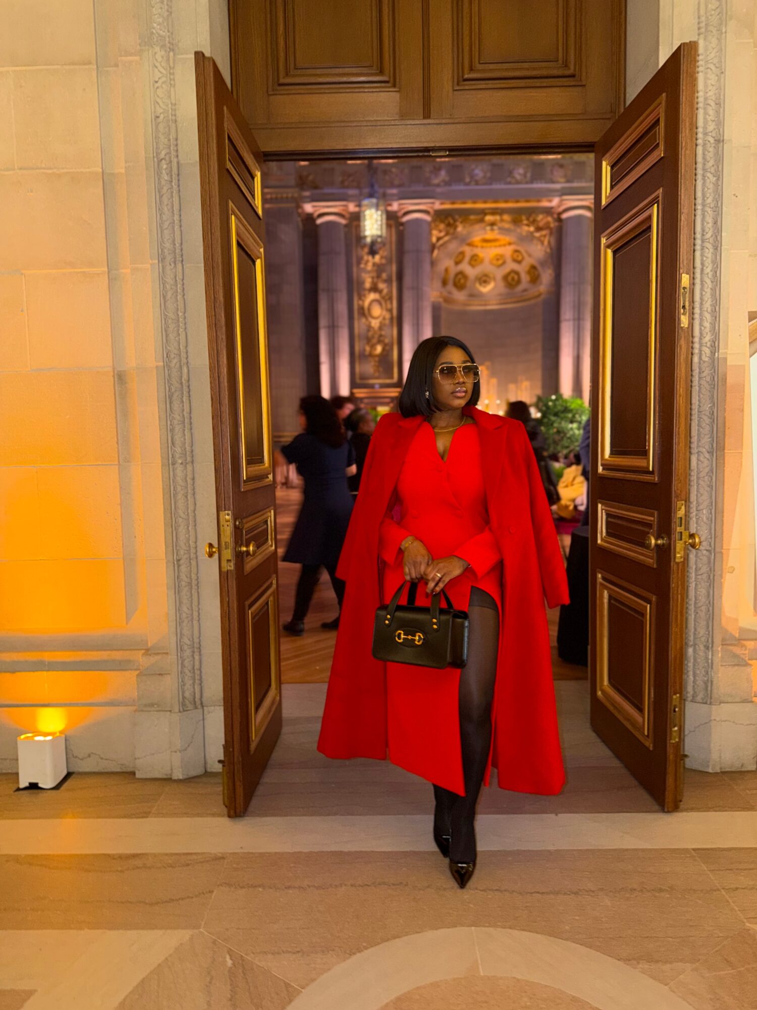 Woman in red coat holding a handbag standing in doorway with ornate interior background.