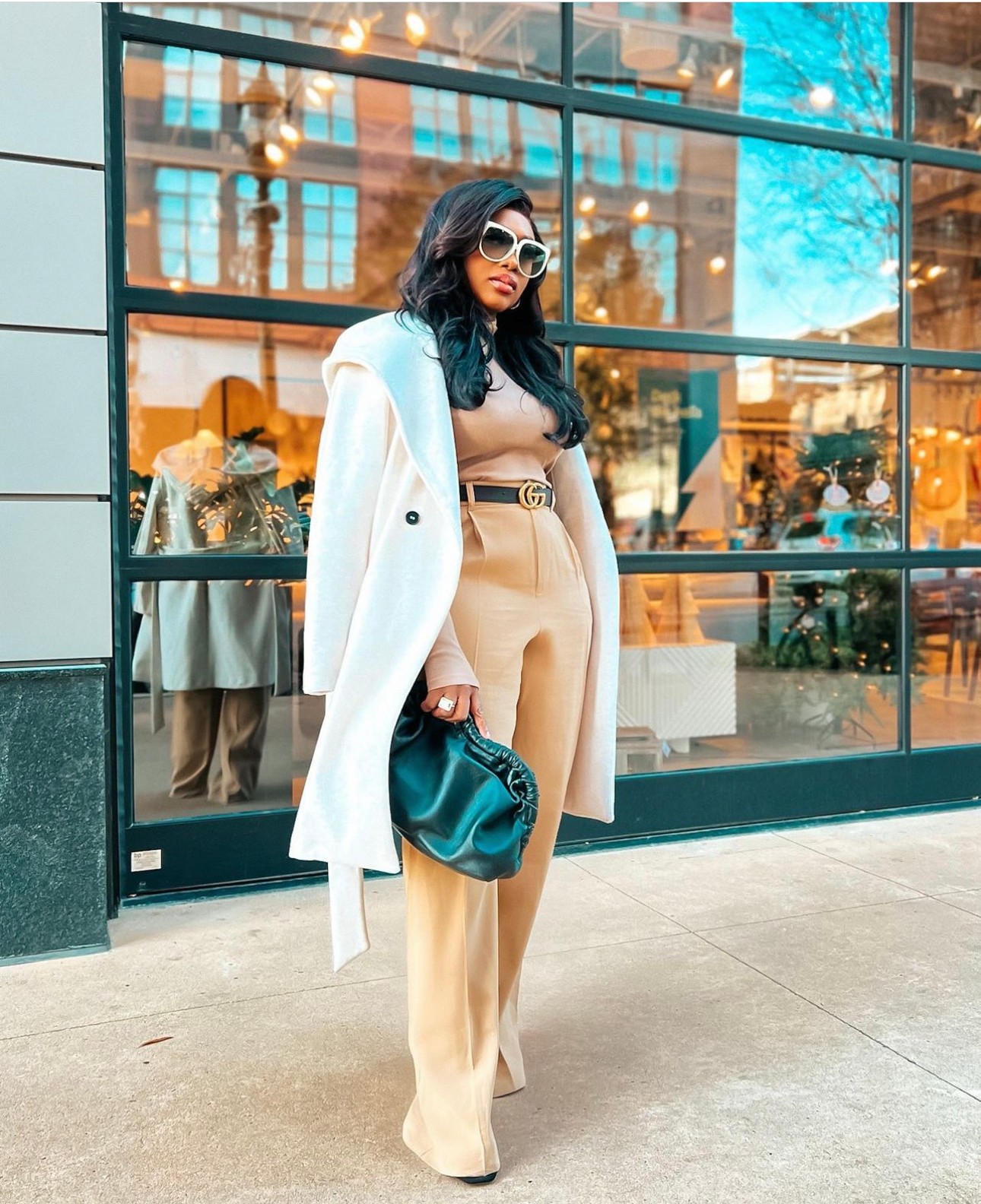 Woman with dark hair and sunglasses standing outside a glass building, wearing a beige coat and pants, holding a blue bag.