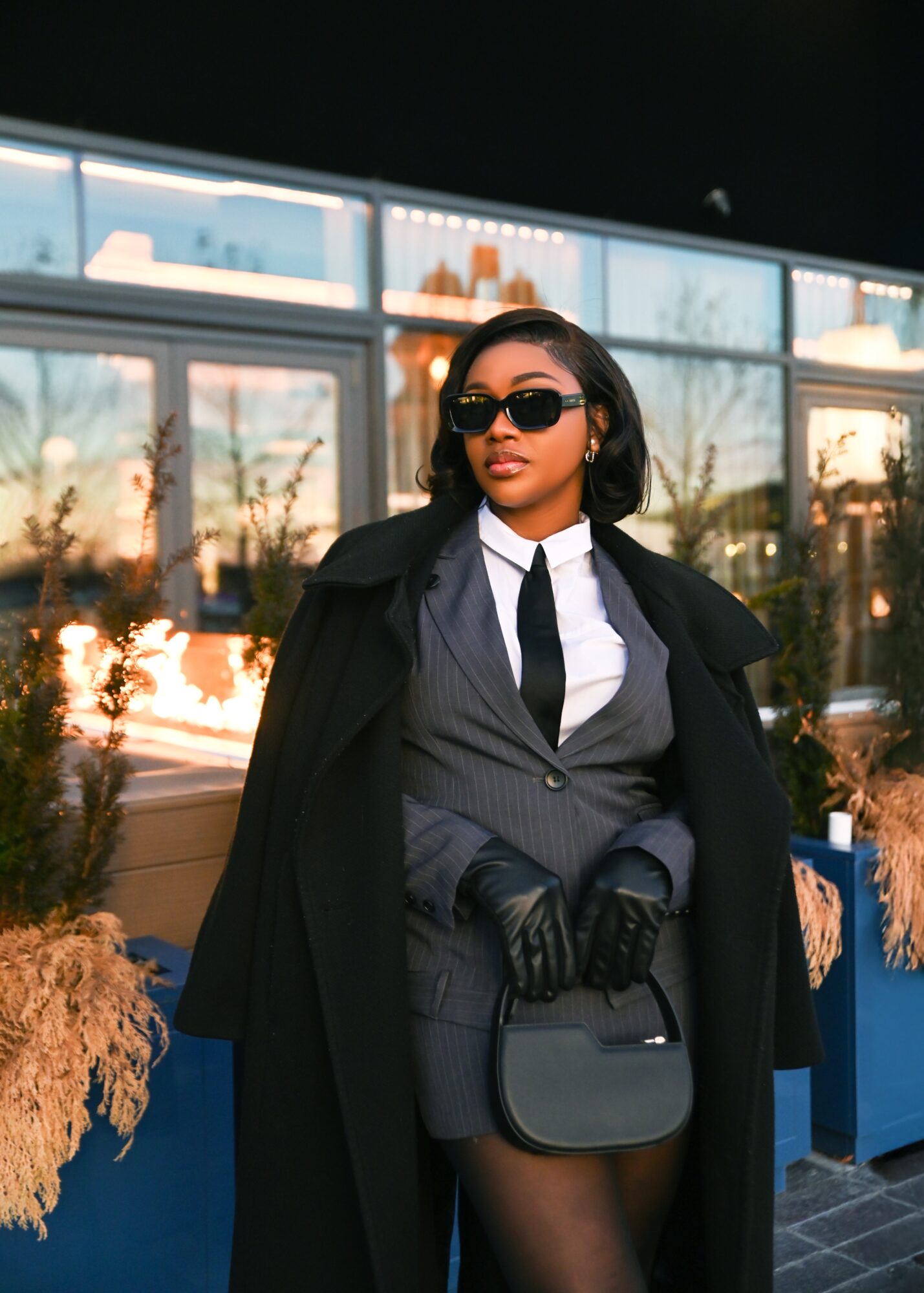 Woman in sunglasses, suit, and coat standing outdoors with buildings and trees behind her.