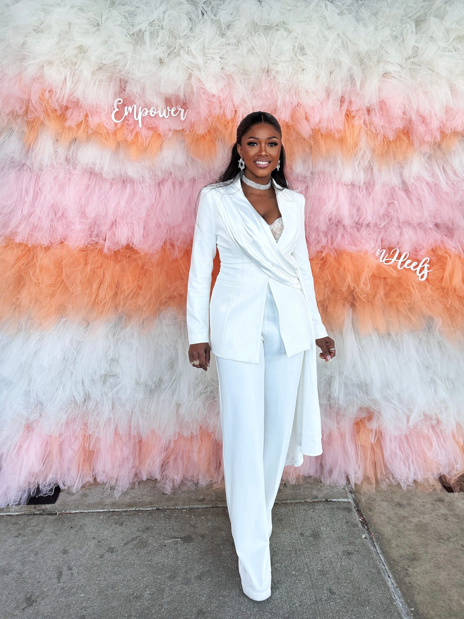 Woman in white suit standing in front of colorful feather backdrop with words 'Empower' and 'Hearts'.