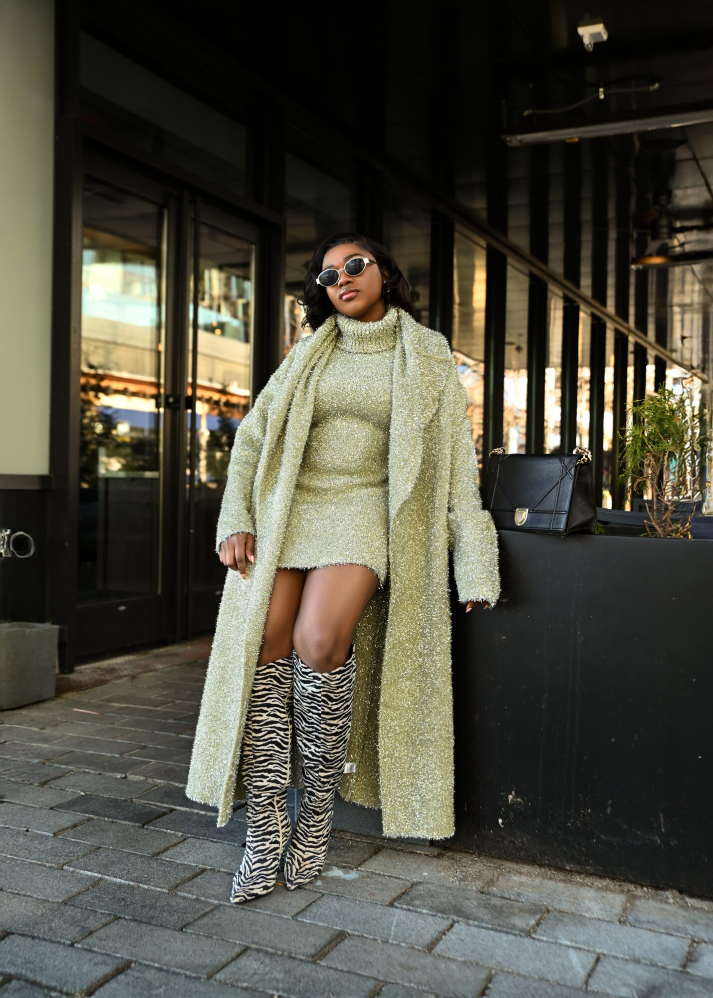 Woman in a cozy coat and sunglasses standing outside a building with glass windows and a black counter, wearing zebra-print boots.